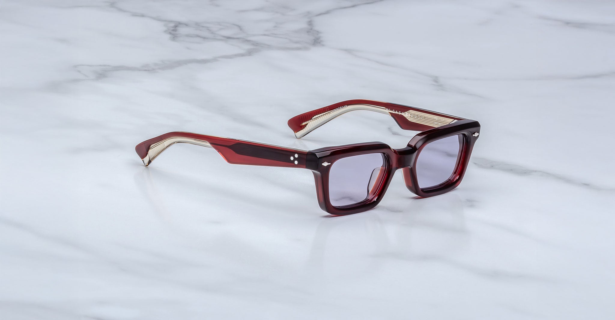 The Jacques Marie Mage Casablanca eyeglasses feature a rectangular, thick frame in dark red with clear accents, tinted lenses, and gold-toned arm details, displayed on a white marble surface.
