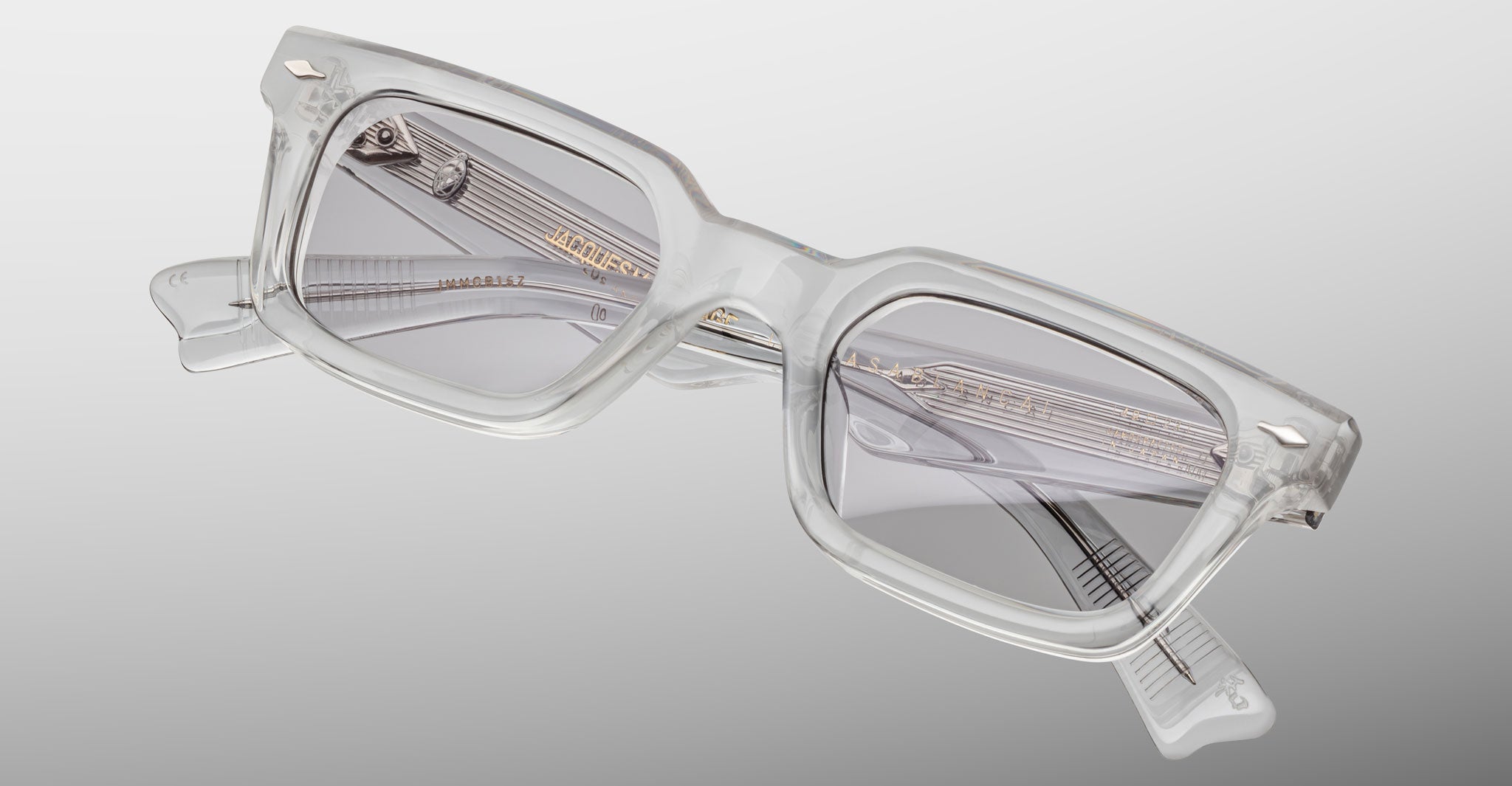 The Jacques Marie Mage Casablanca features clear acetate rectangular frames with light gray tinted lenses, visible hardware near the hinges, and gold text on the inner arm, shown against a gradient gray background.