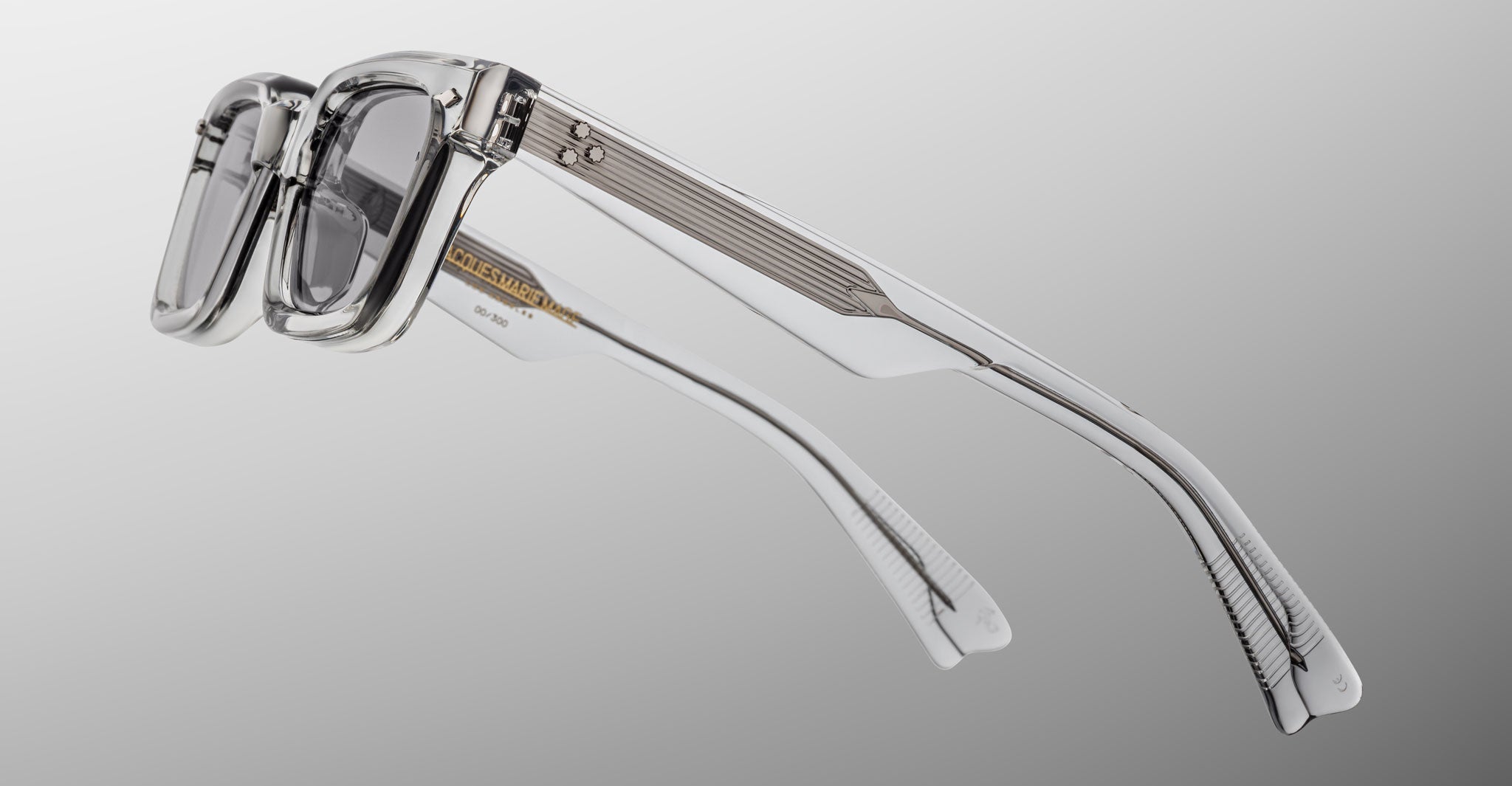 Jacques Marie Mage Casablanca eyeglasses feature clear frames, grey-tinted lenses, gold accents, and the brand name on the inner left arm, set against a light gradient background.