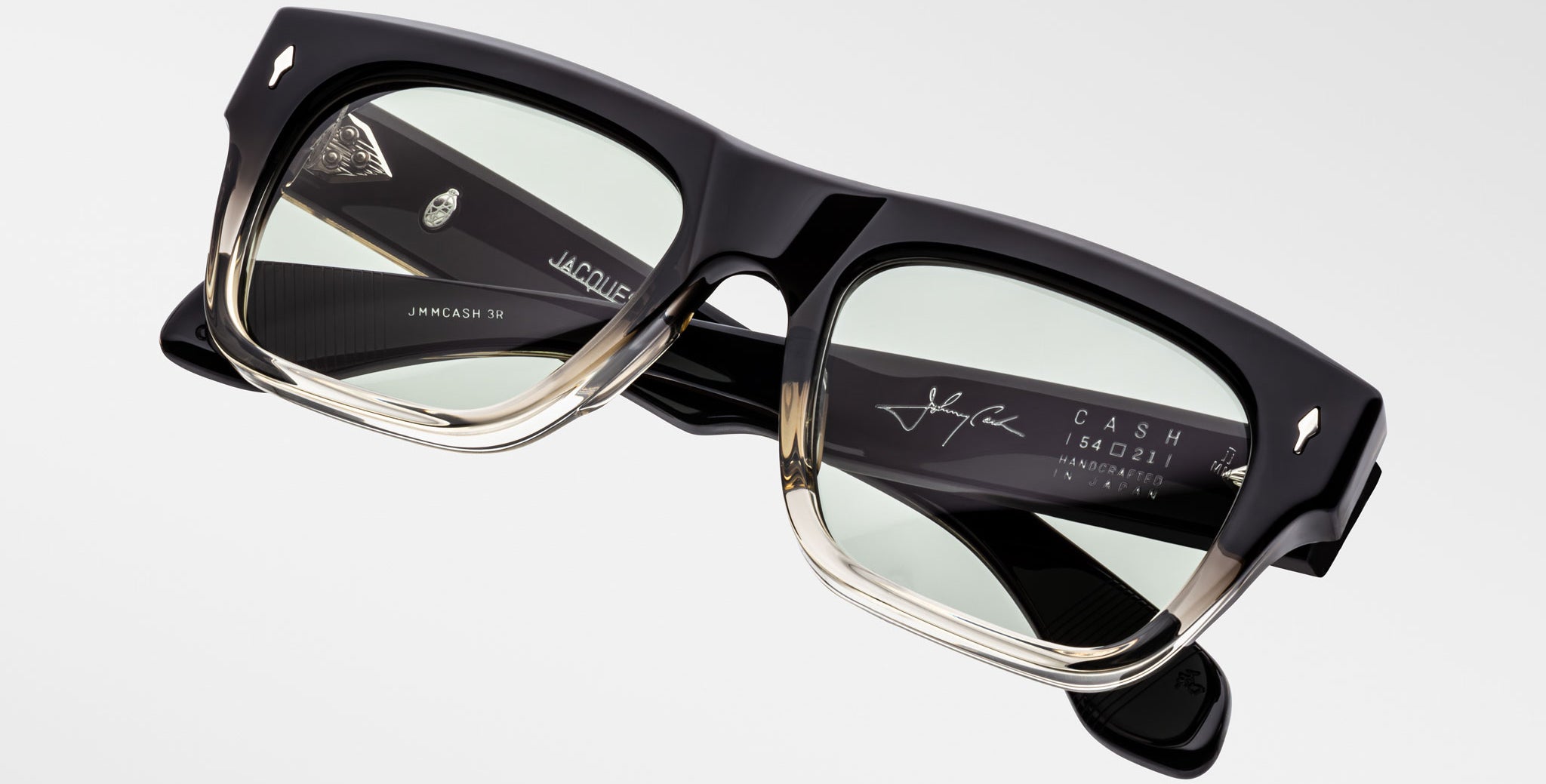 A close-up shows the Jacques Marie Mage Cash rectangular black and clear acetate eyeglasses with green-tinted lenses, highlighting engraved logos, text, and a signature on the inside of the temples.