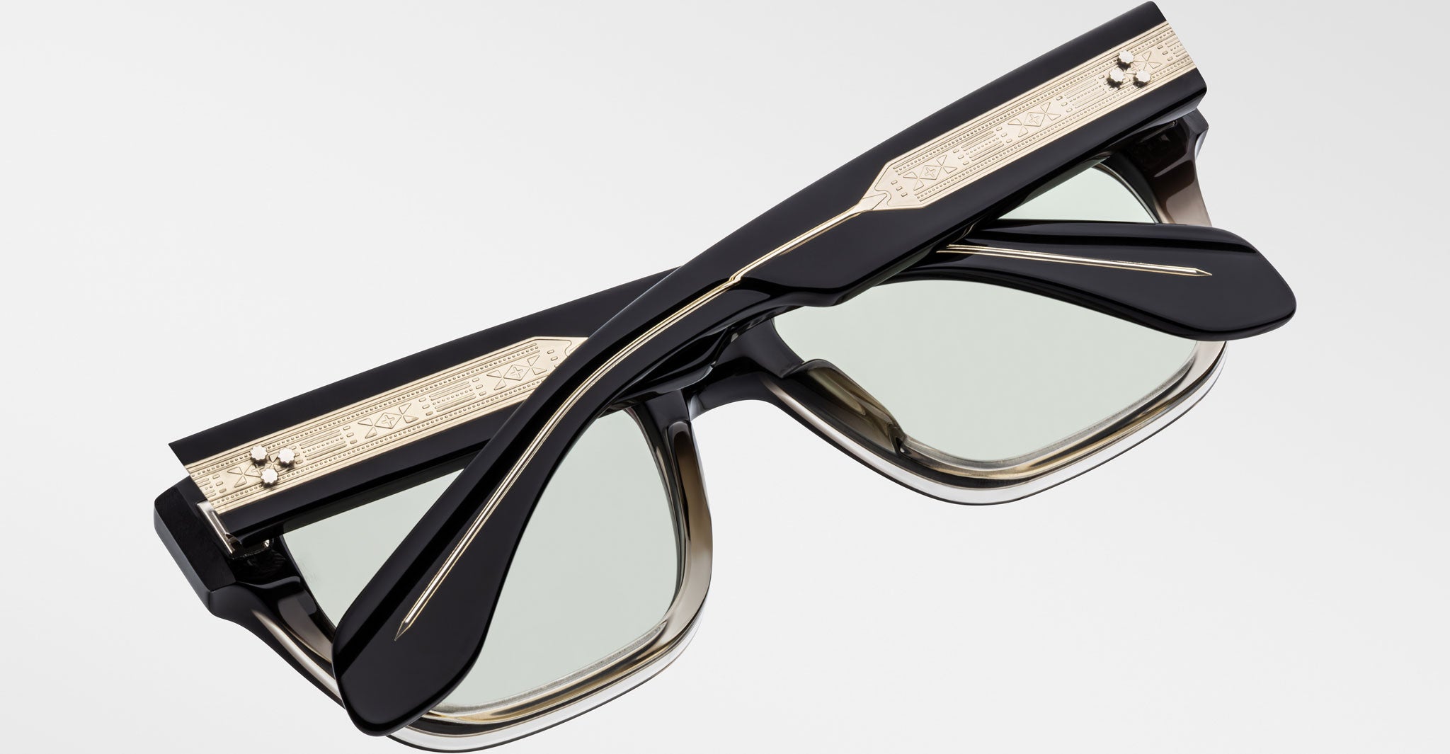 The Jacques Marie Mage Cash eyeglasses are black and rectangular with lightly tinted lenses, folded to display ornate gold temples featuring engraved patterns, all set against a plain white background.