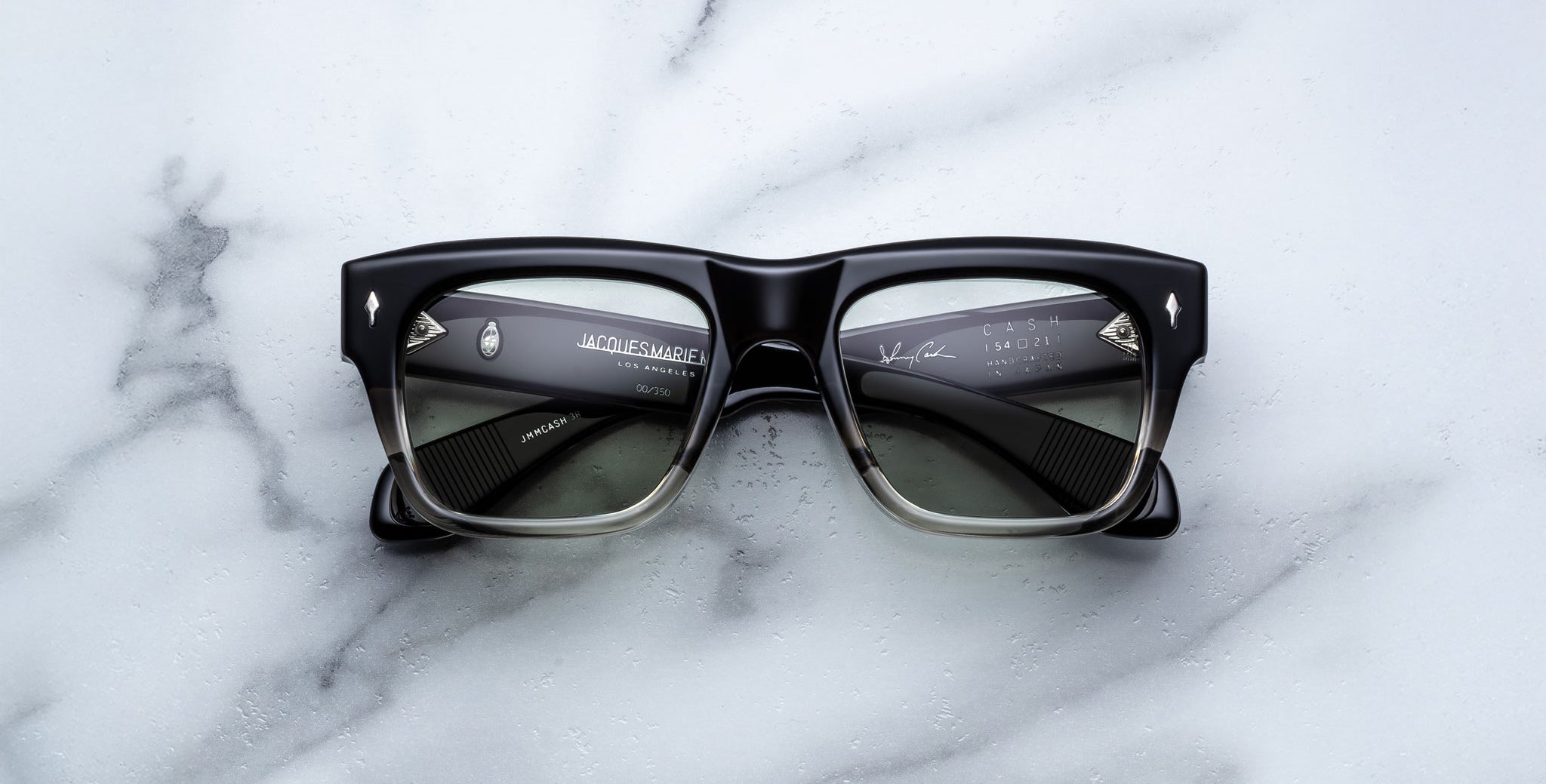 A pair of Jacques Marie Mage Cash black rectangular sunglasses with slightly translucent lenses rests on a white marble surface; the inner arms display branding and product details in white text.