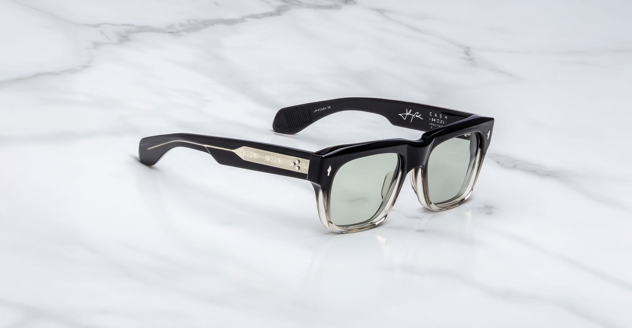 The Jacques Marie Mage Cash sunglasses, featuring a black square frame with metallic temple accents and lightly tinted lenses, are shown on a white marble surface.
