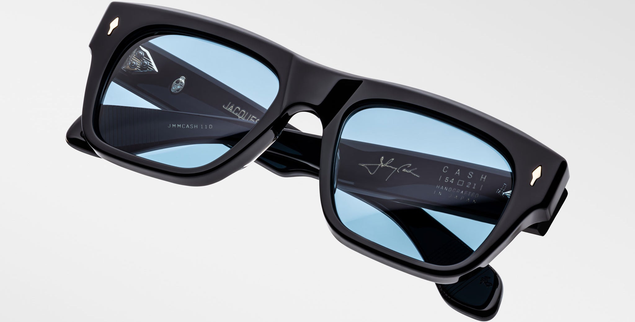 A pair of Jacques Marie Mage Cash black rectangular sunglasses with light blue tinted lenses is displayed against a plain background. The inner arms show white and gold branding, signature, and product details.
