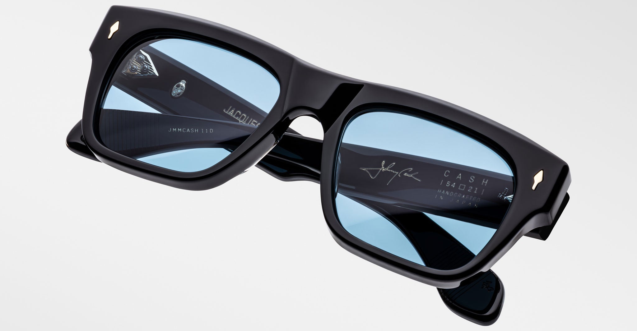 A pair of Jacques Marie Mage Cash black rectangular sunglasses with light blue tinted lenses is displayed against a plain background. The inner arms show white and gold branding, signature, and product details.
