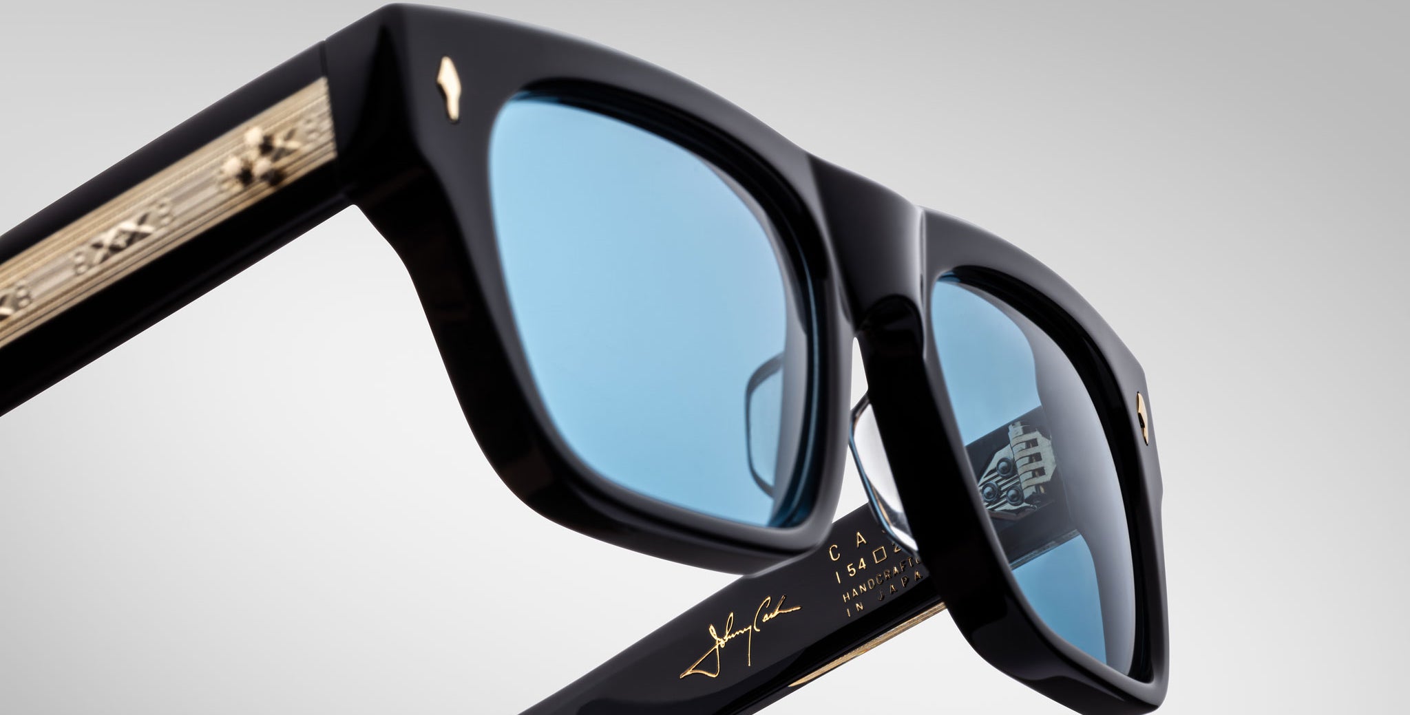 Close-up view of Jacques Marie Mage Cash black sunglasses with blue-tinted lenses, gold detailing on the arms, and a signature on the inner frame, photographed against a plain light background.