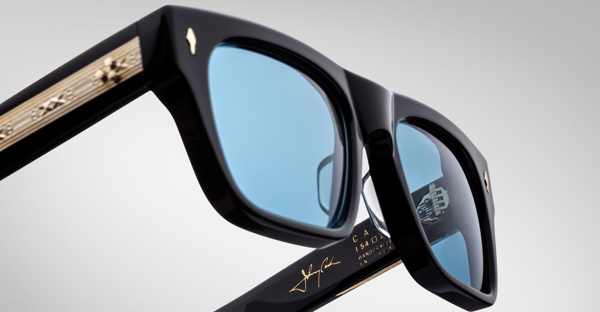 Close-up view of Jacques Marie Mage Cash black sunglasses with blue-tinted lenses, gold detailing on the arms, and a signature on the inner frame, photographed against a plain light background.