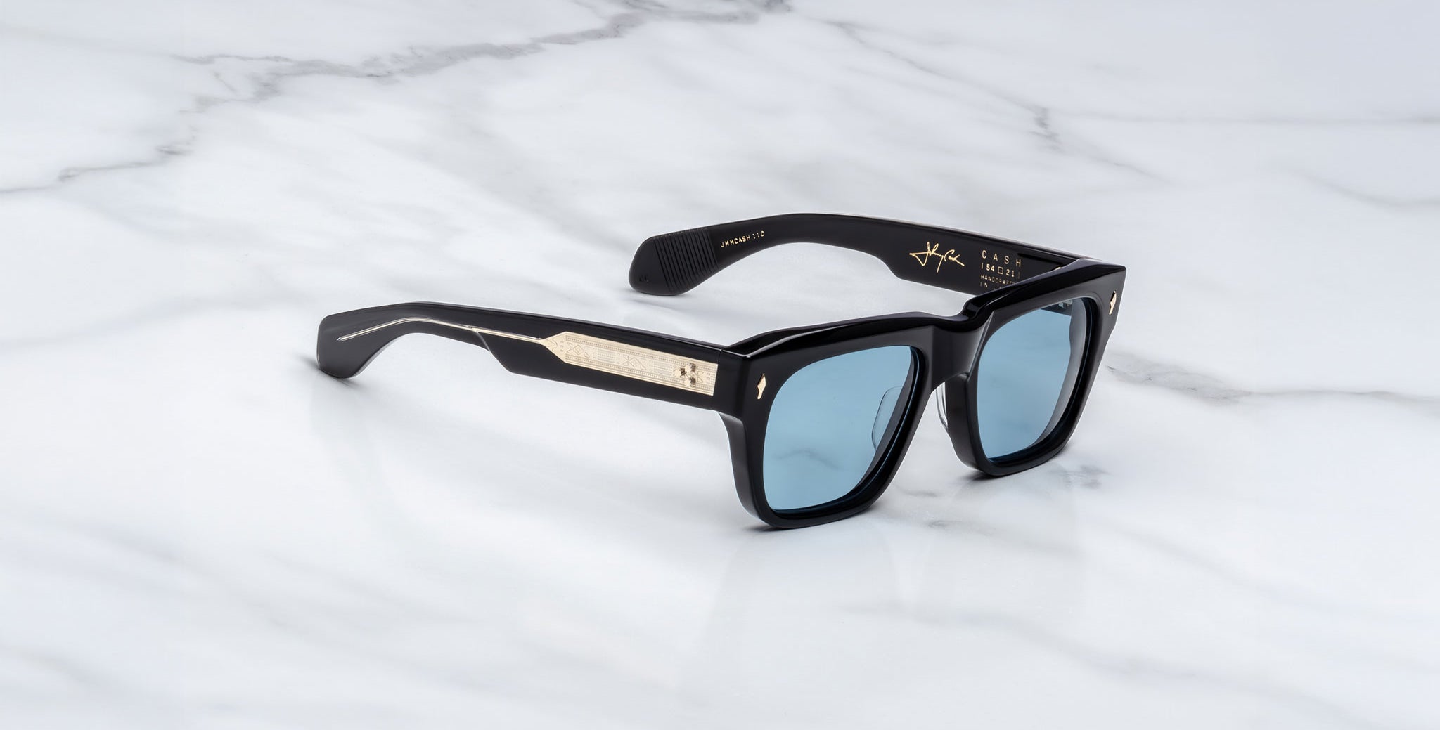 Jacques Marie Mage Cash black rectangular sunglasses with thick frames, blue-tinted lenses, and gold arm detailing are displayed on a white marble surface.