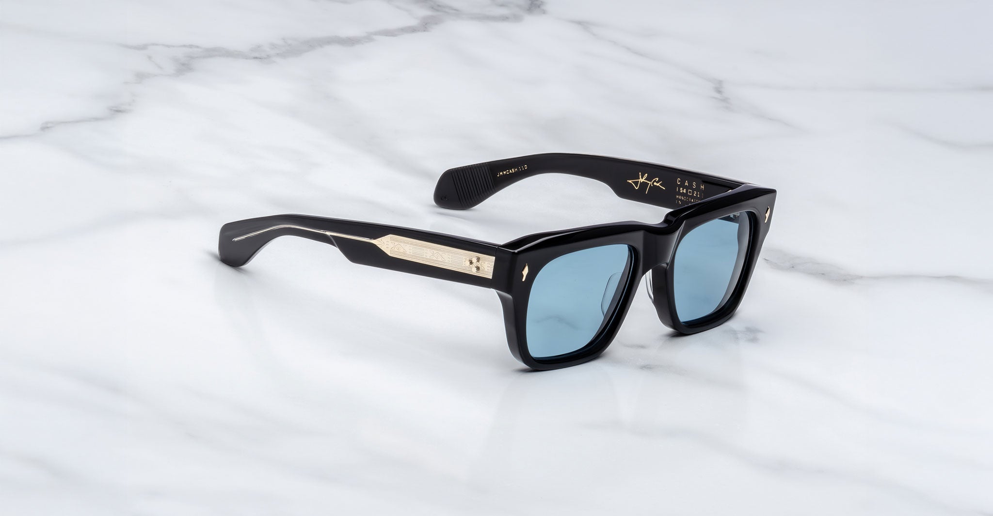 Jacques Marie Mage Cash black rectangular sunglasses with thick frames, blue-tinted lenses, and gold arm detailing are displayed on a white marble surface.
