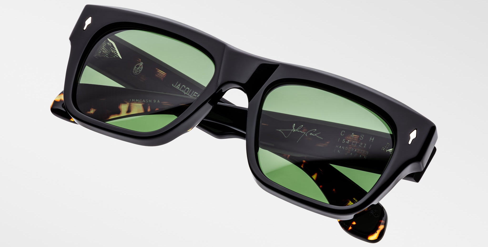 The Jacques Marie Mage Cash sunglasses feature black rectangular frames, green lenses, and tortoiseshell-detailed arms with branded and etched signatures on the inner sides, shown against a plain light background.