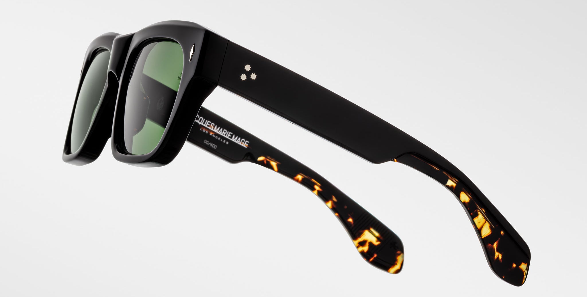The Jacques Marie Mage Cash sunglasses feature thick black frames, green-tinted lenses, two small circular logos on one temple, and tortoiseshell detailing on the lower arms.