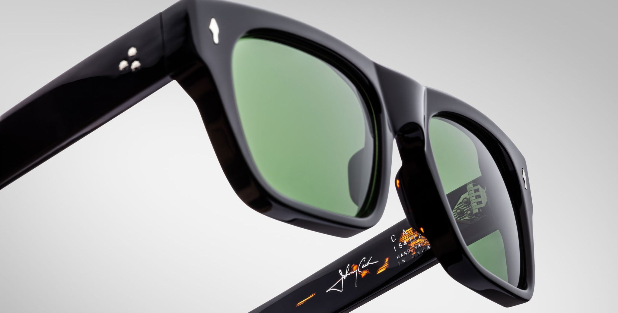 Close-up view of Jacques Marie Mage Cash black sunglasses with green-tinted lenses, silver stud accents on the temples, and orange and white signature-style markings on the inner arm, set against a plain light gray background.