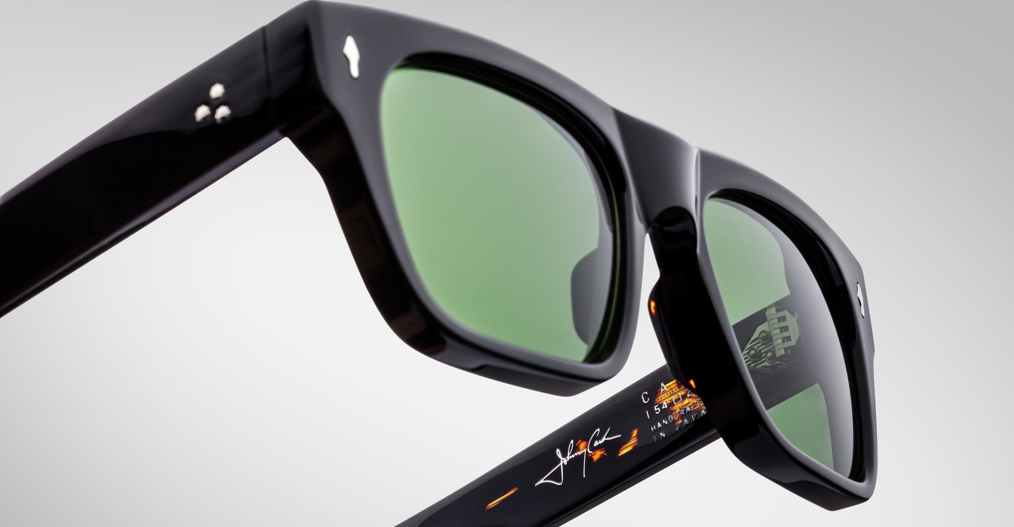 Close-up view of Jacques Marie Mage Cash black sunglasses with green-tinted lenses, silver stud accents on the temples, and orange and white signature-style markings on the inner arm, set against a plain light gray background.