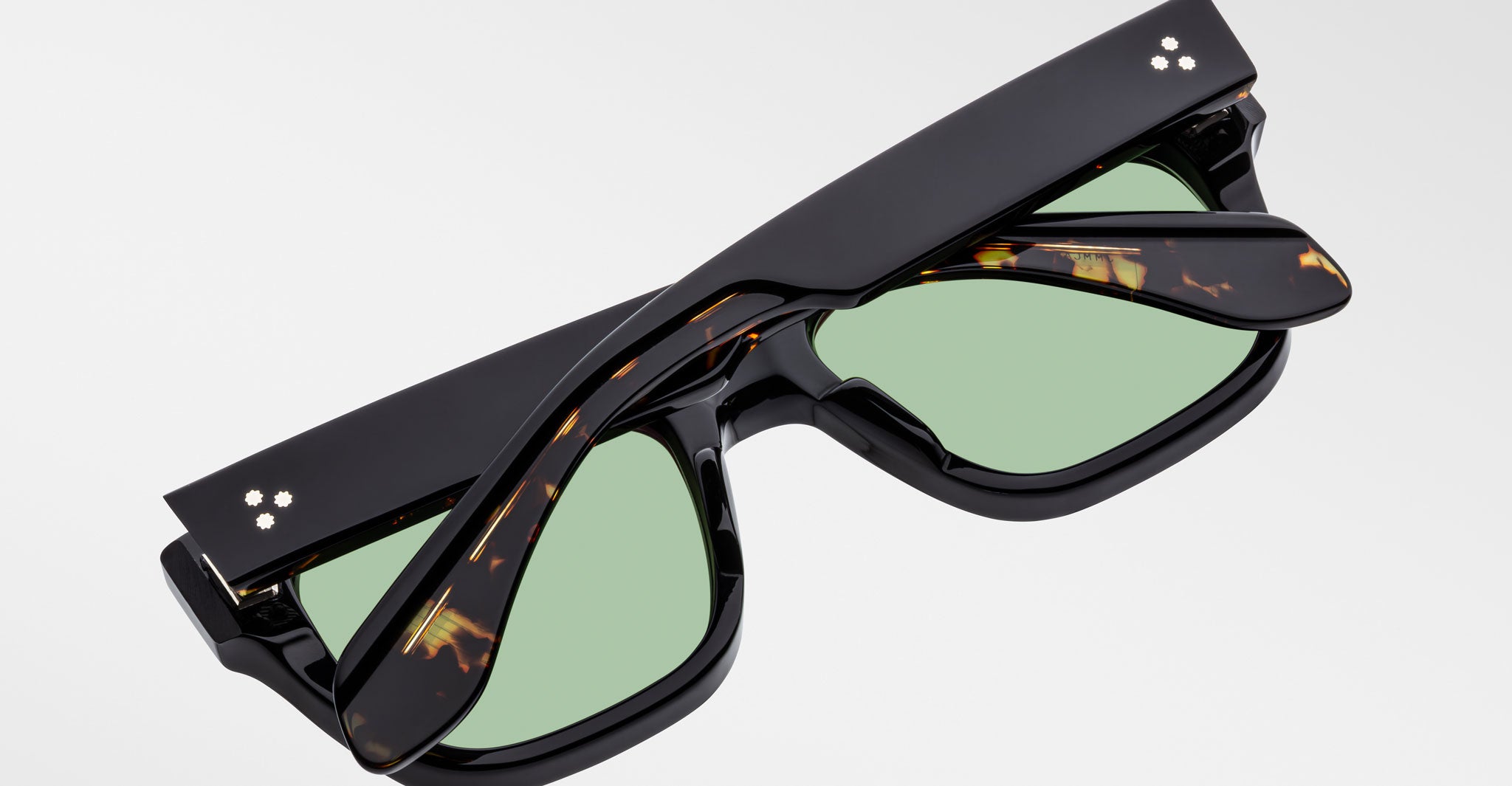 A folded pair of Jacques Marie Mage Cash black sunglasses with green lenses and tortoiseshell-patterned inner arms rests on a plain white surface.