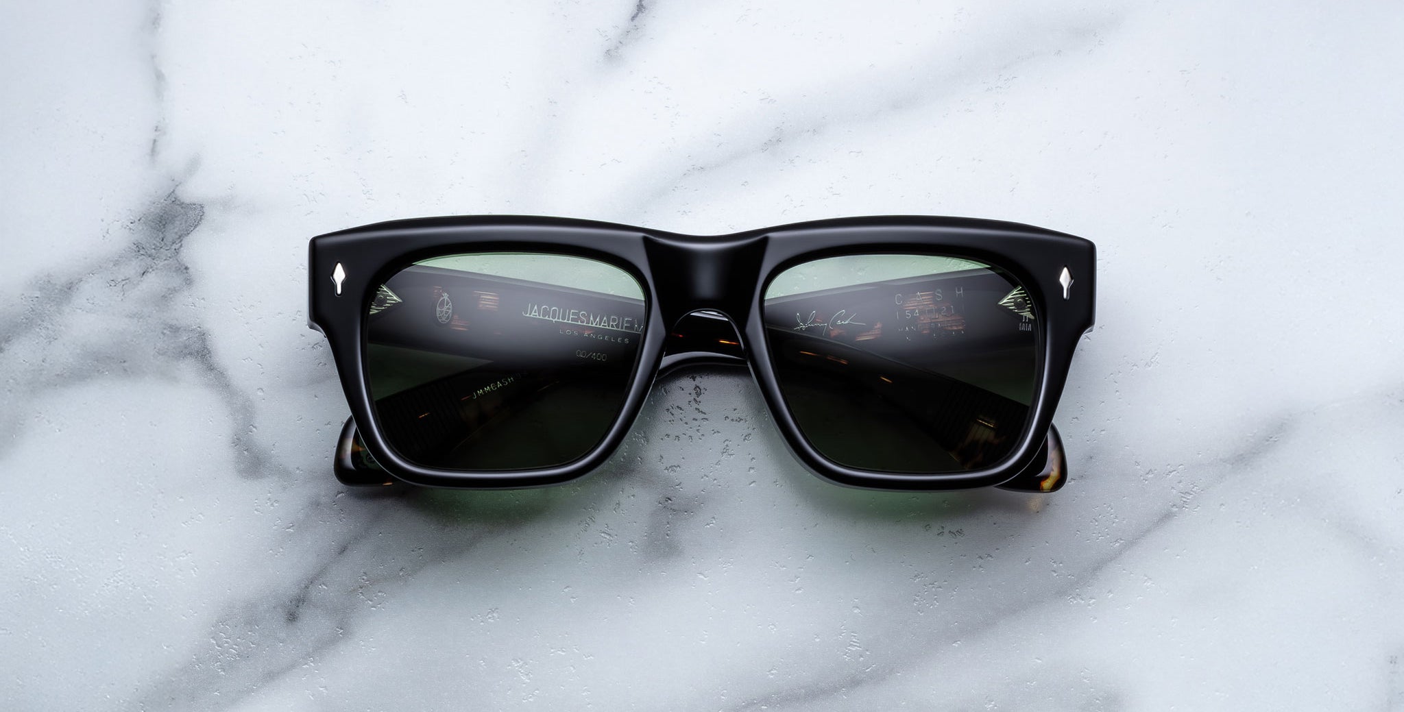 A pair of Jacques Marie Mage Cash black rectangular sunglasses with dark lenses rests on a white marble surface, reflecting dashboard instruments on the lenses.