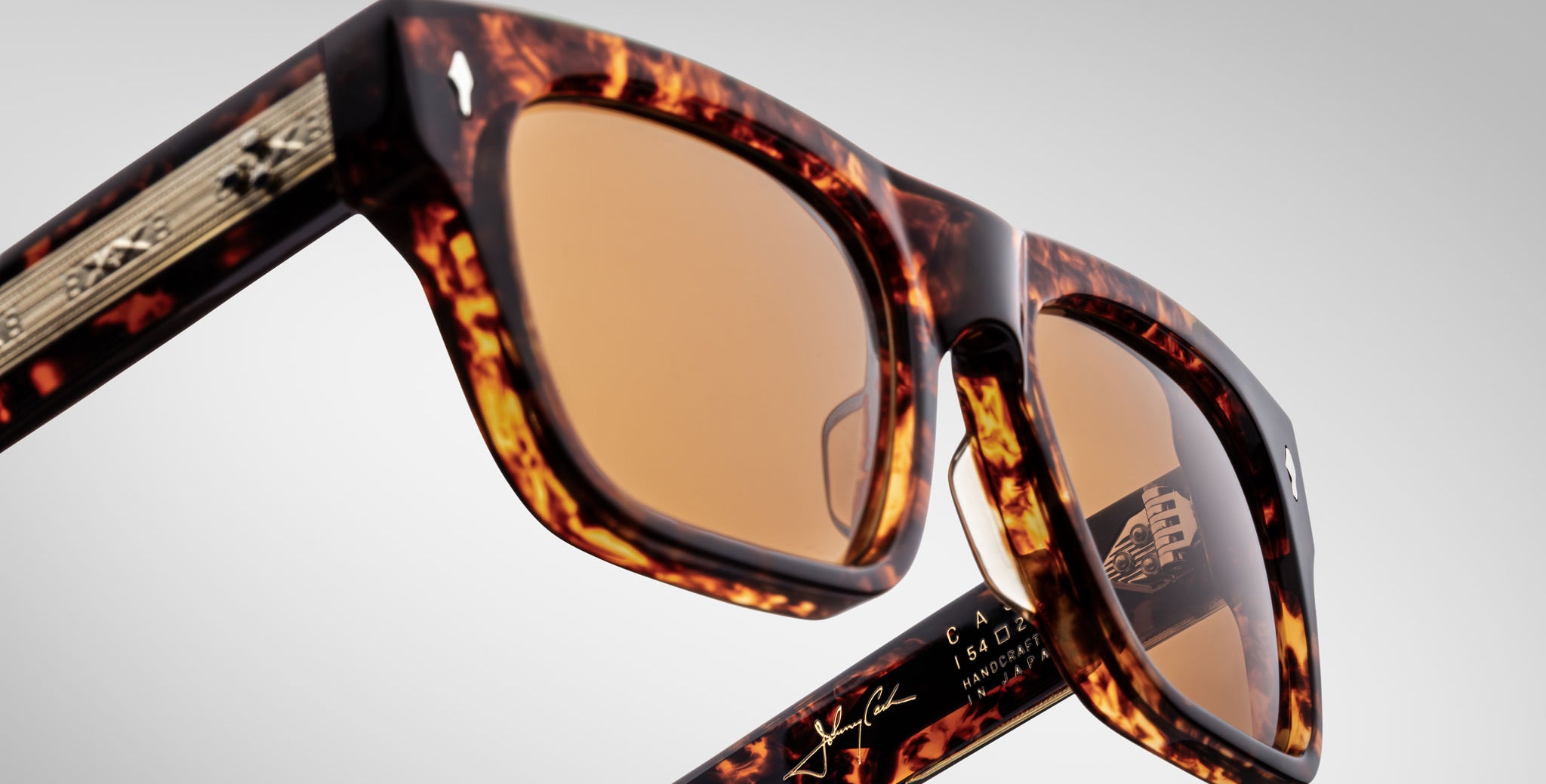 Close-up of Jacques Marie Mage Cash sunglasses with tortoiseshell frame and amber lenses, shown against a plain background. Features gold detailing on the arms plus visible branding and signatures inside the arms.