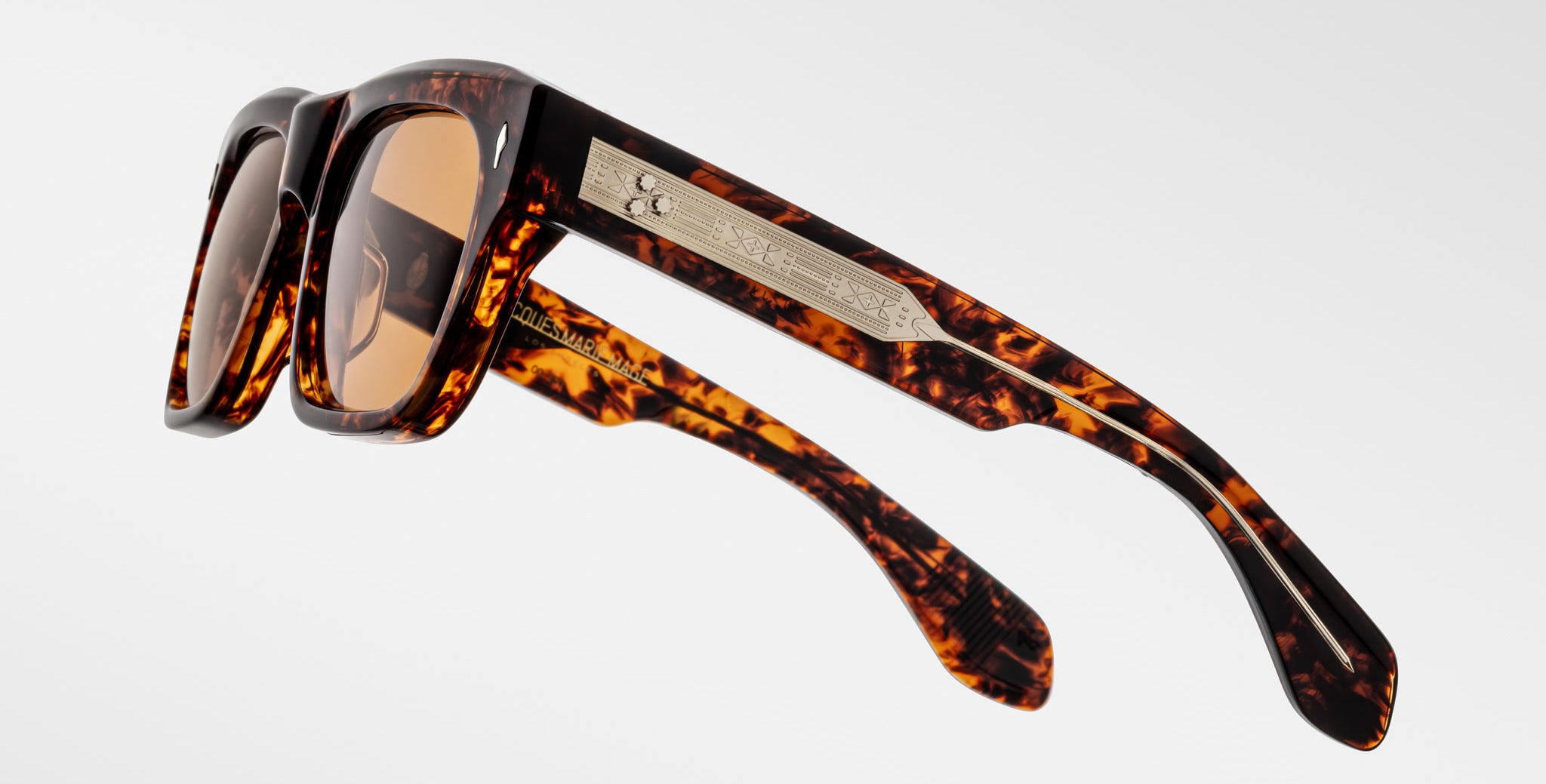 A close-up of Jacques Marie Mage Cash sunglasses featuring tortoiseshell-patterned rectangular frames, brown lenses, and arms adorned with a metallic plaque engraved with text and a small emblem, set against a plain white background.