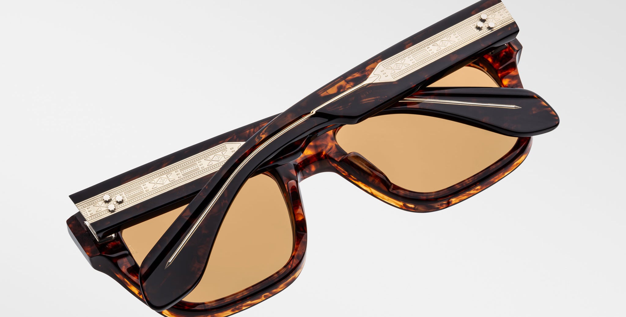 A pair of Jacques Marie Mage Cash tortoiseshell sunglasses with brown lenses, highlighted by gold-toned decorative metal accents on the arms and top edge, is shown folded against a plain light background.