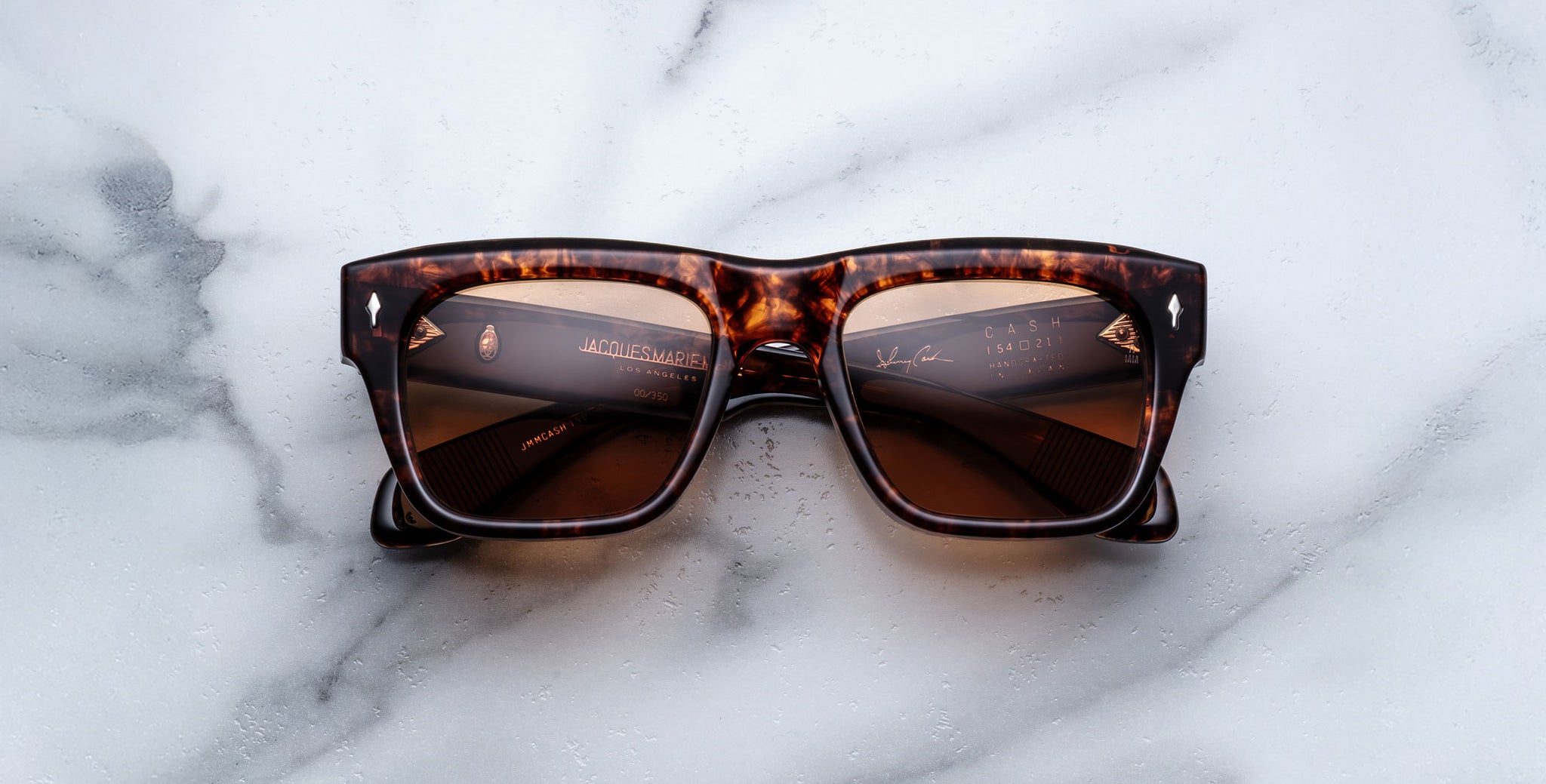 A pair of Jacques Marie Mage Cash rectangular tortoiseshell sunglasses with brown lenses rests on a white marble surface, featuring some text and reflections visible on the lenses.