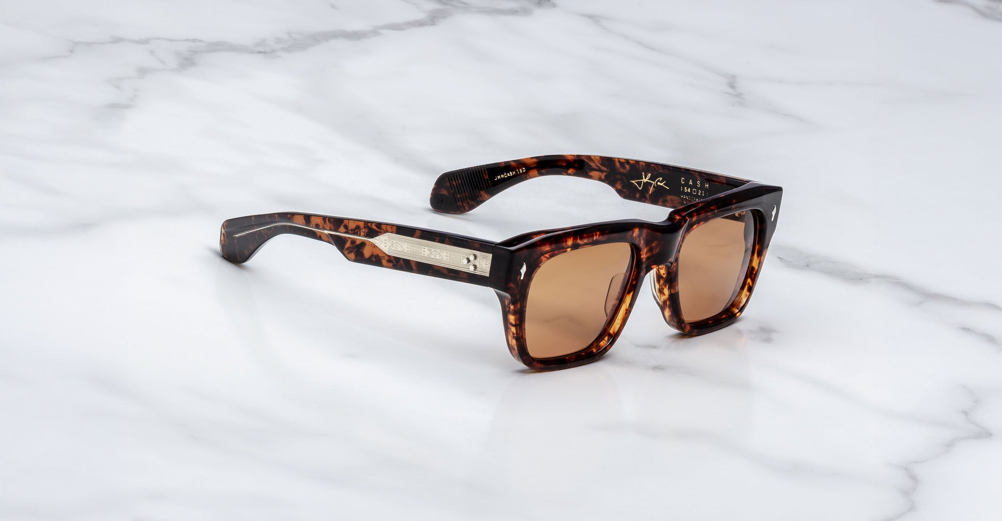 The Jacques Marie Mage Cash sunglasses feature a rectangular tortoiseshell frame, brown lenses, thick arms, and a metallic accent on the temple, shown on a white marble surface.