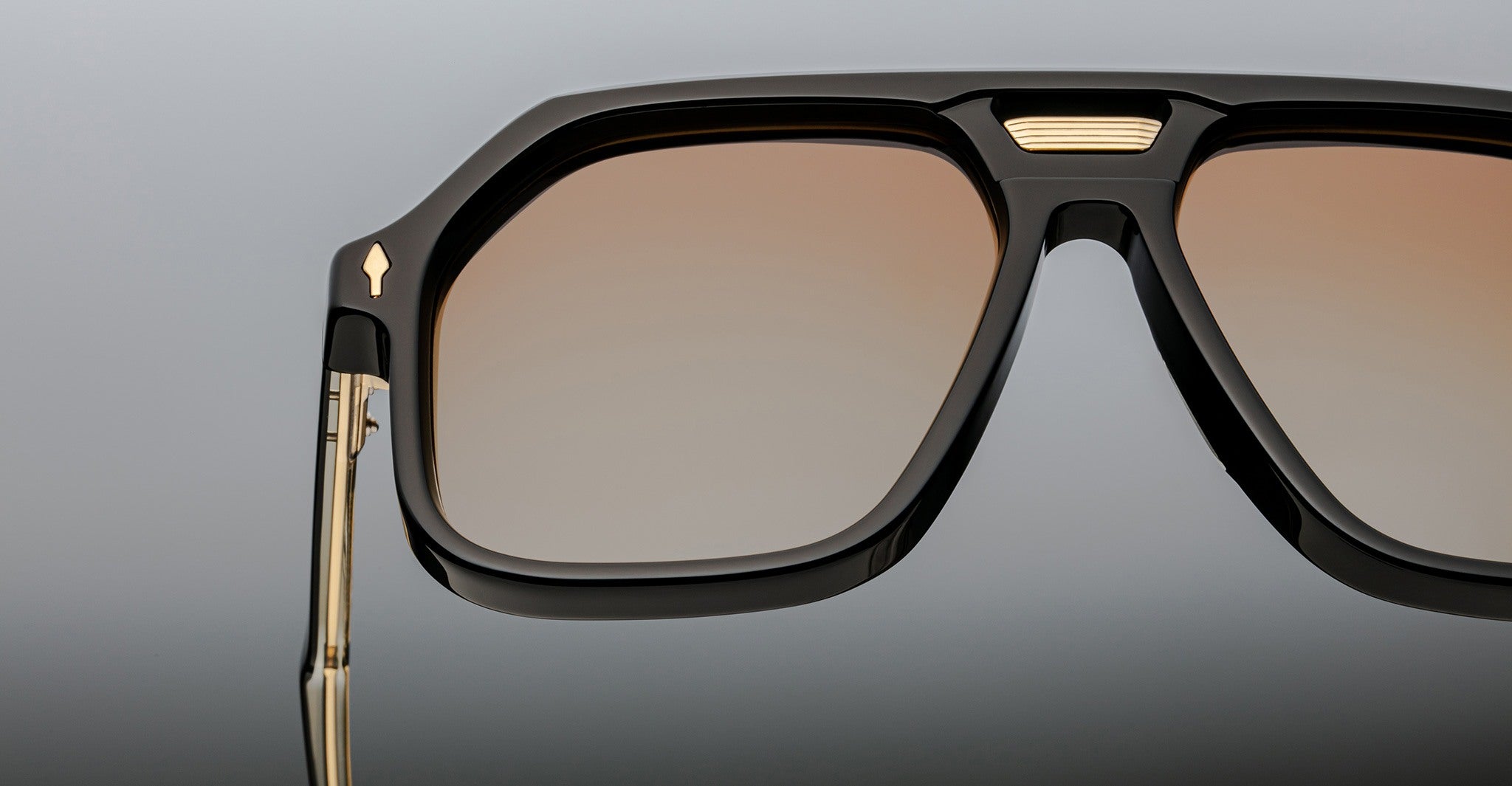 Close-up view of Jacques Marie Mage Casius sunglasses featuring stylish black frames, brown-tinted lenses, gold accents, and a gradient gray background.
