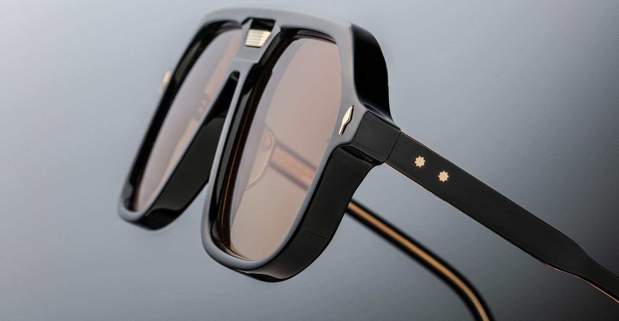 Close-up of Jacques Marie Mage Casius sunglasses—stylish black frames with brown-tinted lenses, gold accents, and two small star details on the temples—set against a neutral background.