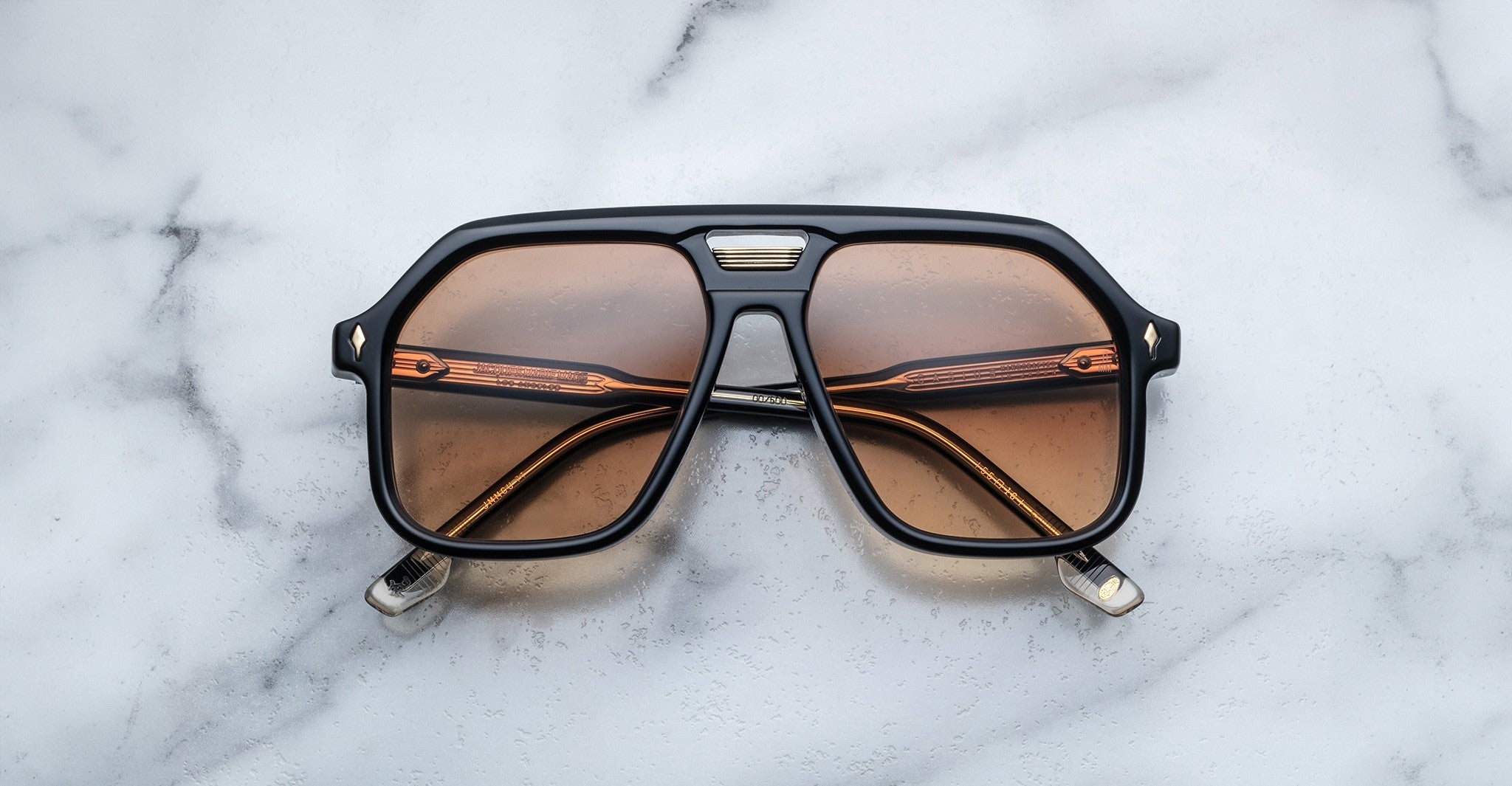 The Jacques Marie Mage Casius, black aviator-style sunglasses with amber-tinted lenses, rests on a white marble surface.
