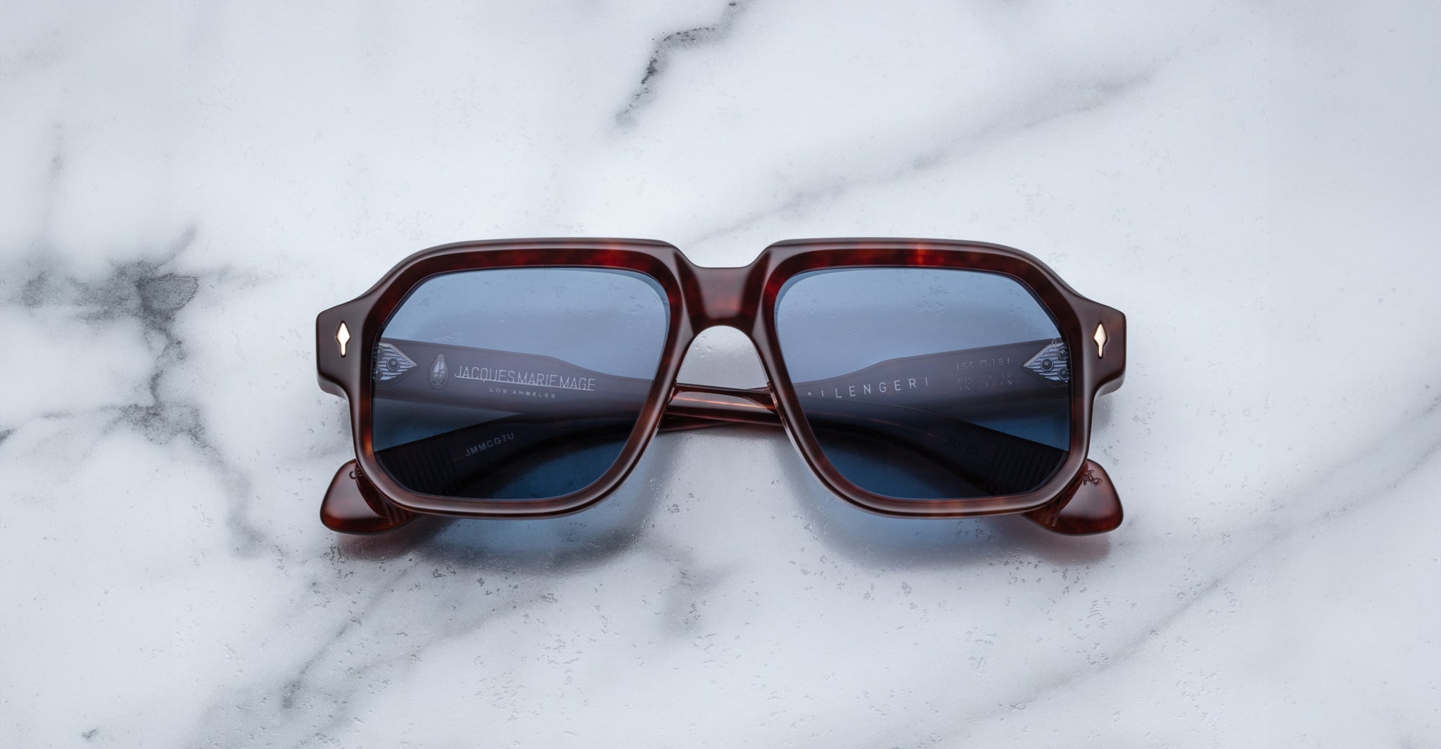 Jacques Marie Mage Challenger sunglasses with square dark tortoiseshell frames and blue-tinted lenses rest on a white marble surface.