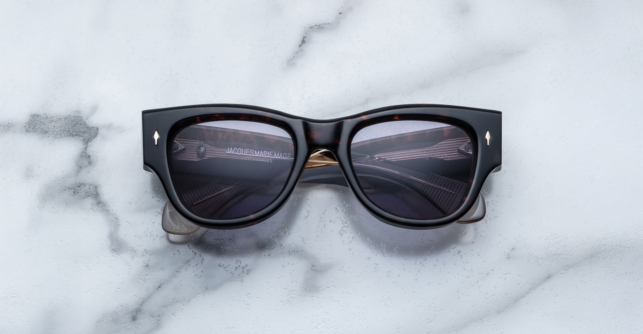 A pair of Jacques Marie Mage Chelsea brown, thick-framed sunglasses with purple lenses rests on a white marble surface with grey veins, with faint brand details visible on the inside of the left lens.