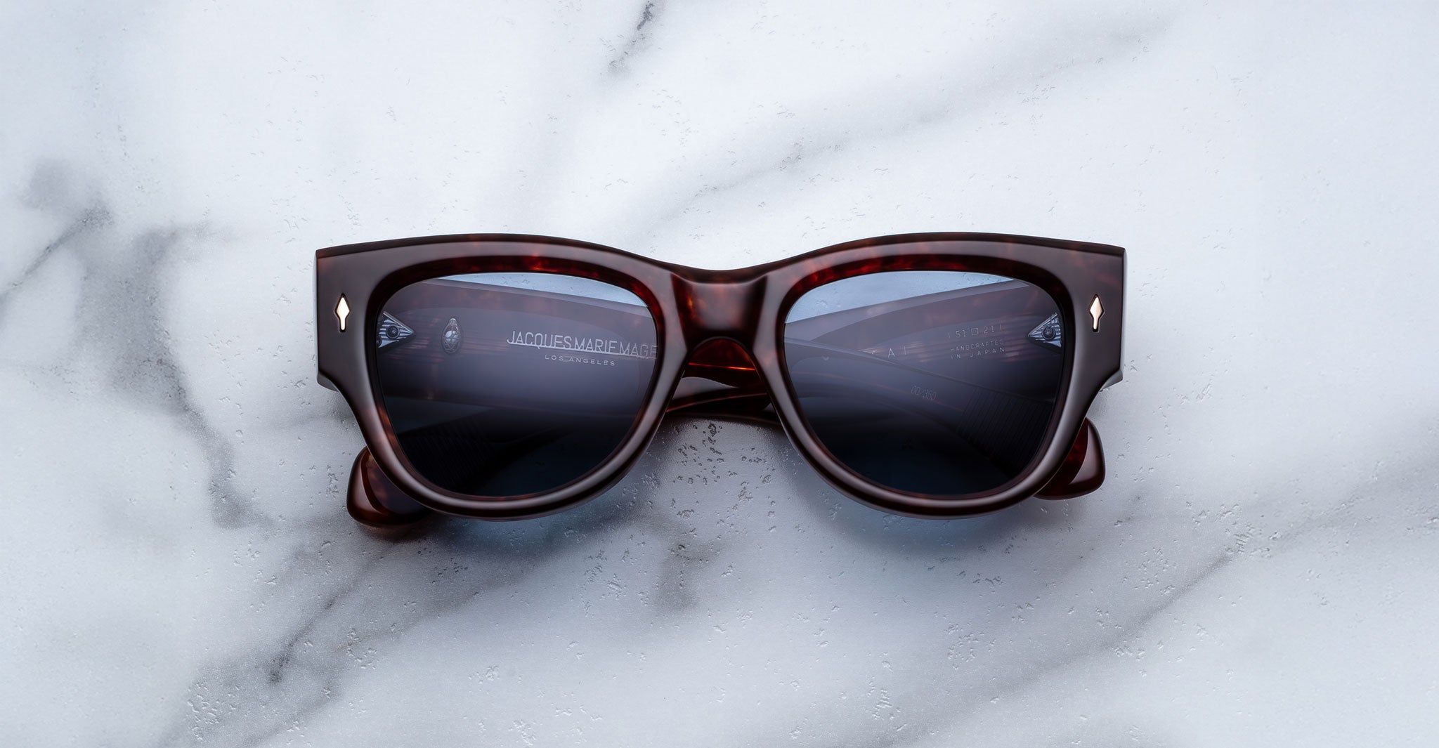 A pair of Jacques Marie Mage Chelsea tortoiseshell sunglasses with black lenses rests on a white marble surface, reflecting a faint image of a computer screen and keyboard.