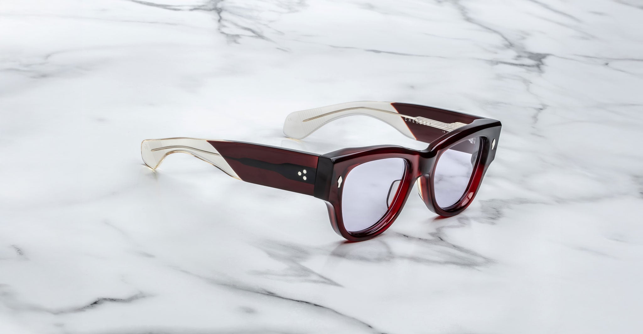 A pair of Jacques Marie Mage Chelsea cherry red, thick-framed sunglasses with light purple lenses sits on a white marble surface with grey veins