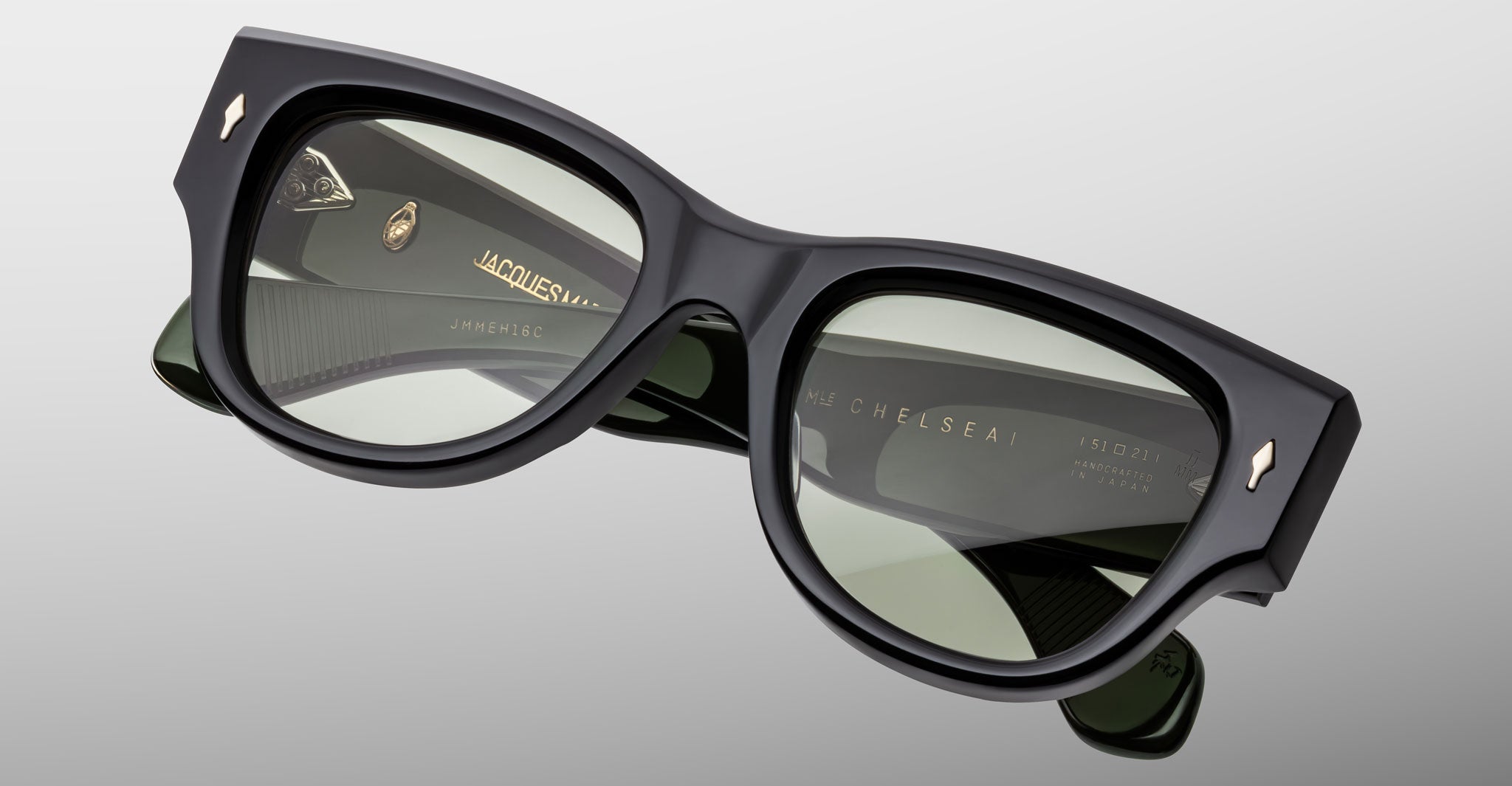 The Jacques Marie Mage Chelsea is a pair of dark, rectangular sunglasses with thick black frames and green-tinted lenses, shown close-up on a light gray background. The inner arms feature engraved gold branding and details.