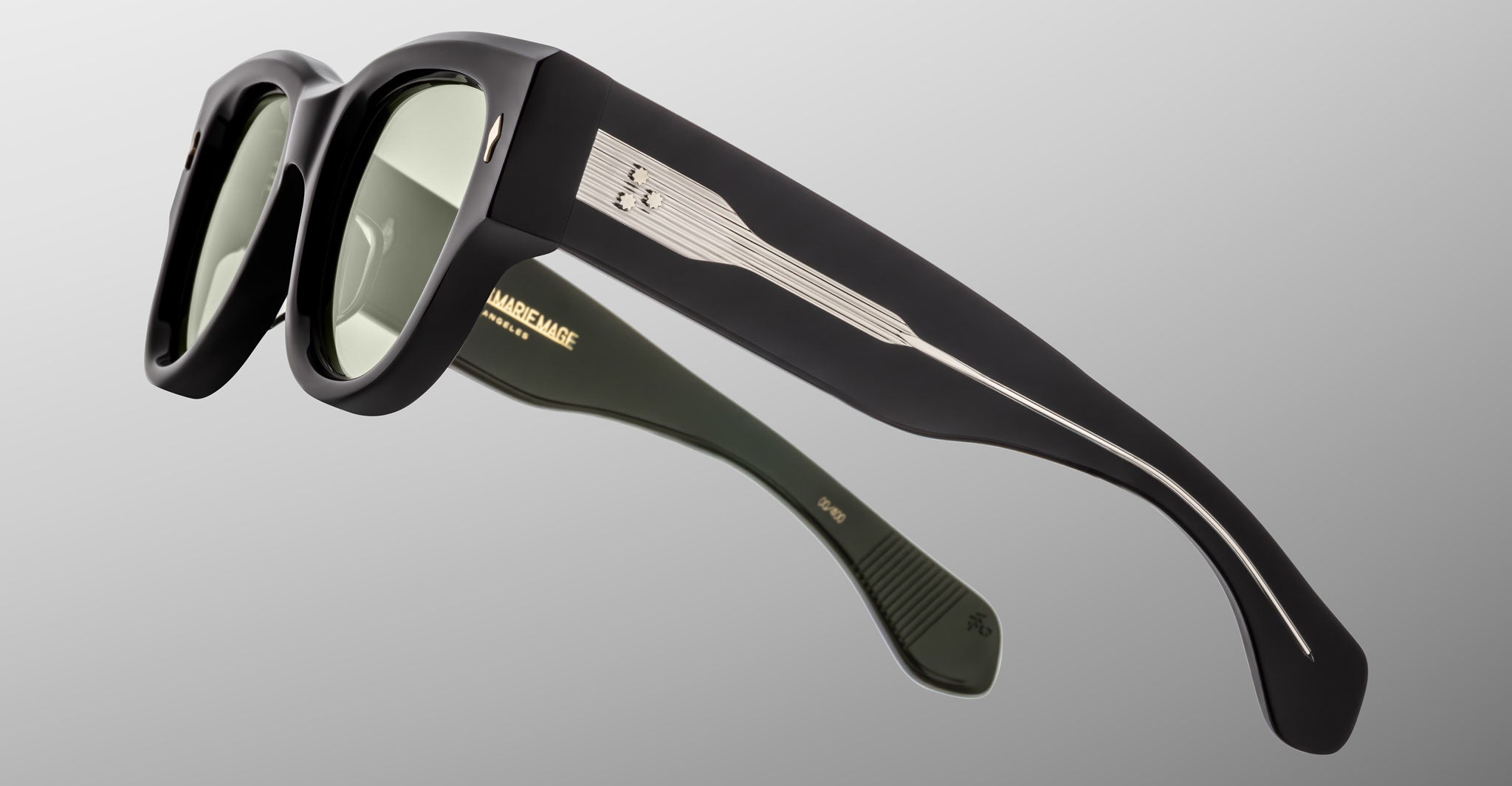 Close-up of Jacques Marie Mage Chelsea sunglasses, featuring thick black frames, green-tinted lenses, and metallic accents on the arms, angled against a plain light gray background.
