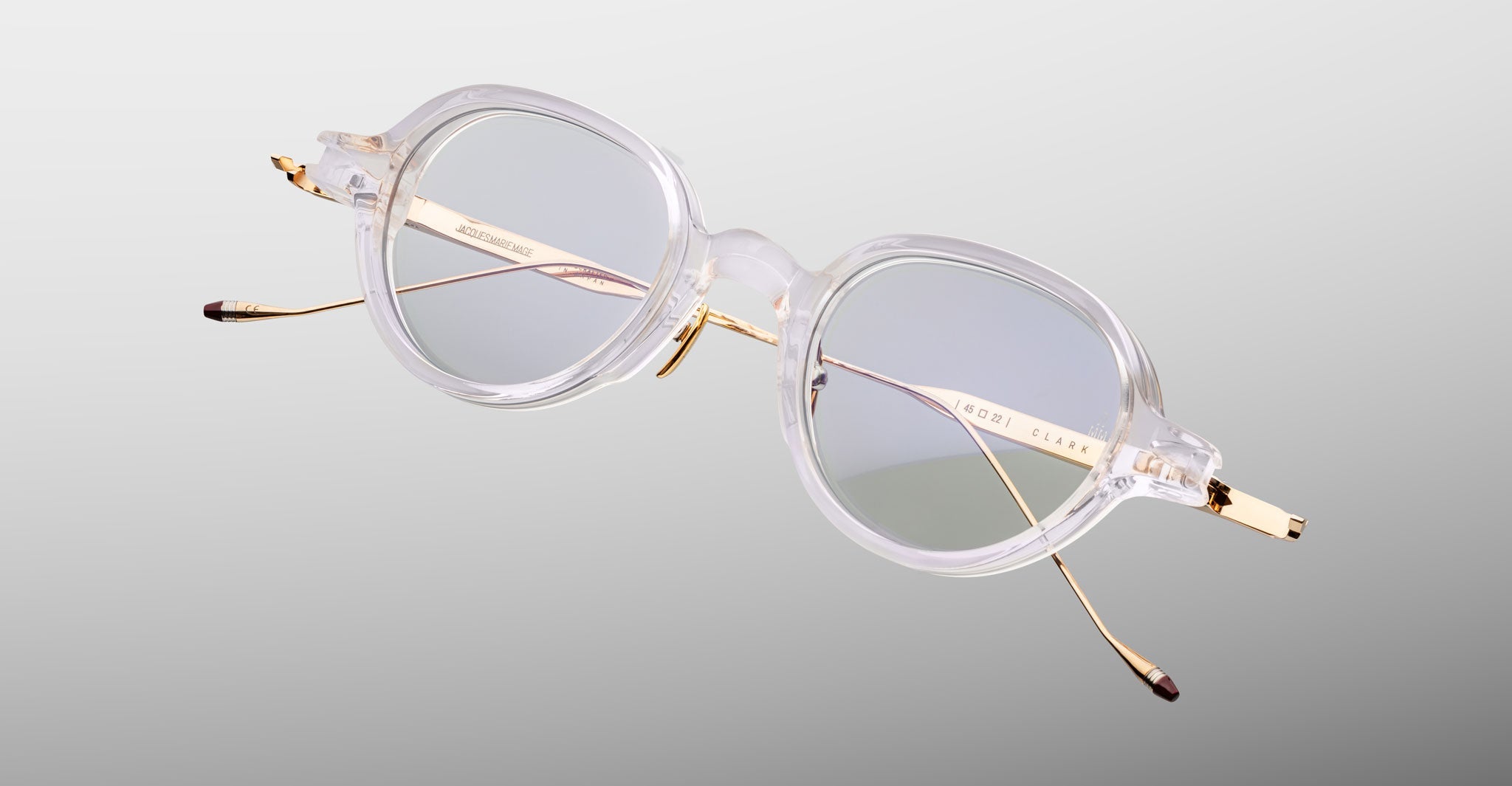 The Jacques Marie Mage Clark eyeglasses feature clear frames, round lenses, and gold metal arms. These midcentury-inspired optical frames by Jacques Marie Mage are shown on a light gray background.