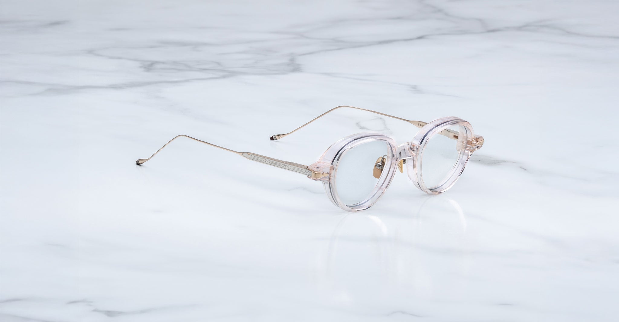 The Jacques Marie Mage Clark features round, clear frames with thin metal arms—midcentury-inspired optical glasses from Jacques Marie Mage—elegantly displayed on a white marble surface.