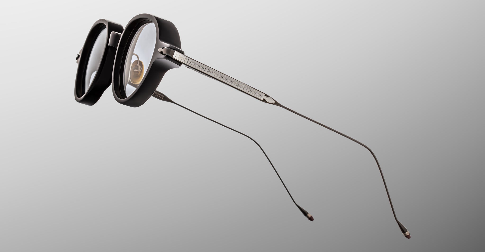 The Jacques Marie Mage Clark by Jacques Marie Mage features black, thick round frames with long, thin metal temples. Handcrafted in Japan and midcentury-inspired, these eyeglasses are displayed against a plain gradient backdrop.