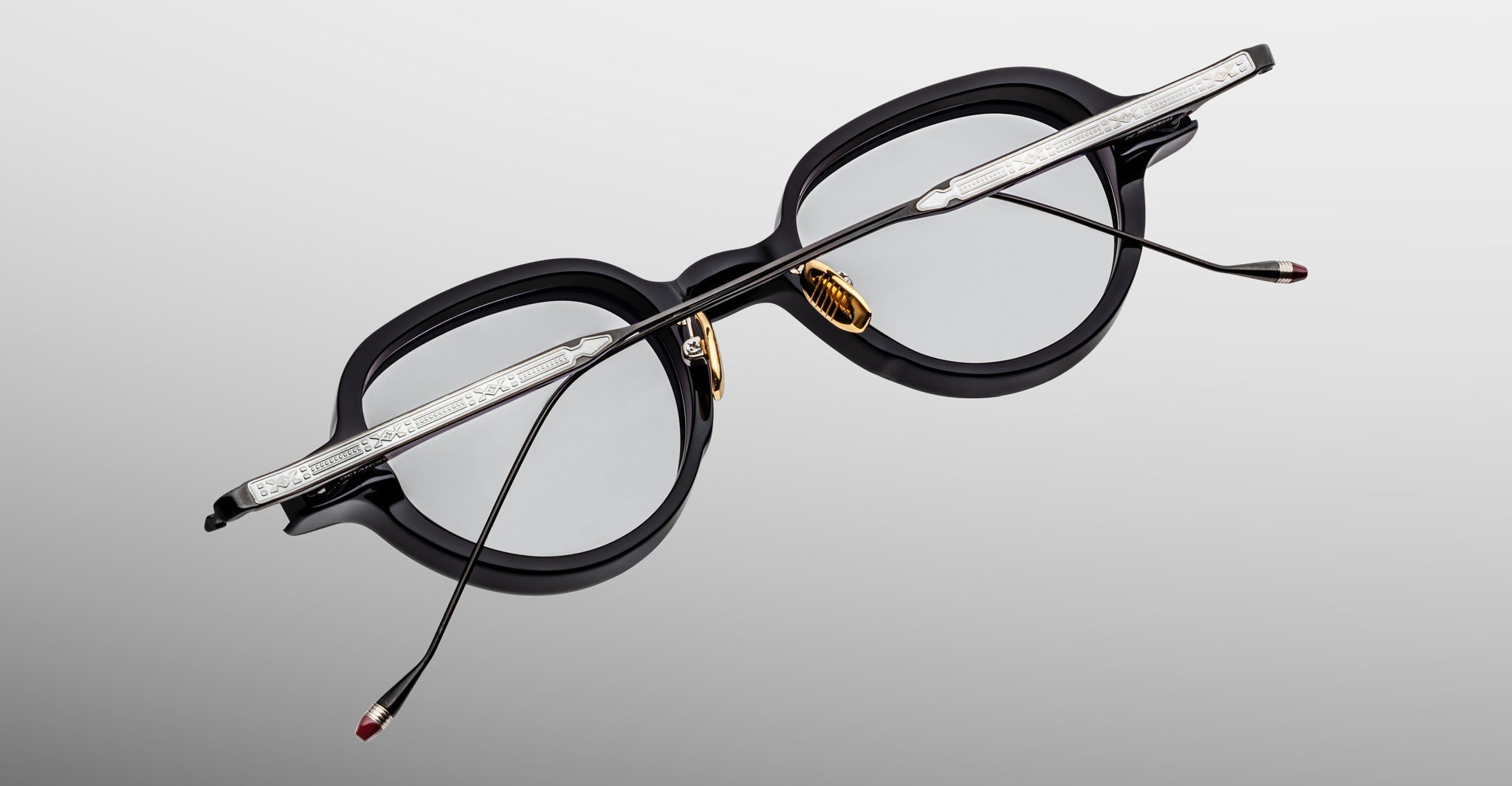 Jacques Marie Mage Clark optical frames feature black rims, silver arms, gold nose pads, and clear lenses. Expertly handcrafted in Japan, these midcentury-inspired glasses by Jacques Marie Mage appear on a light gray background.