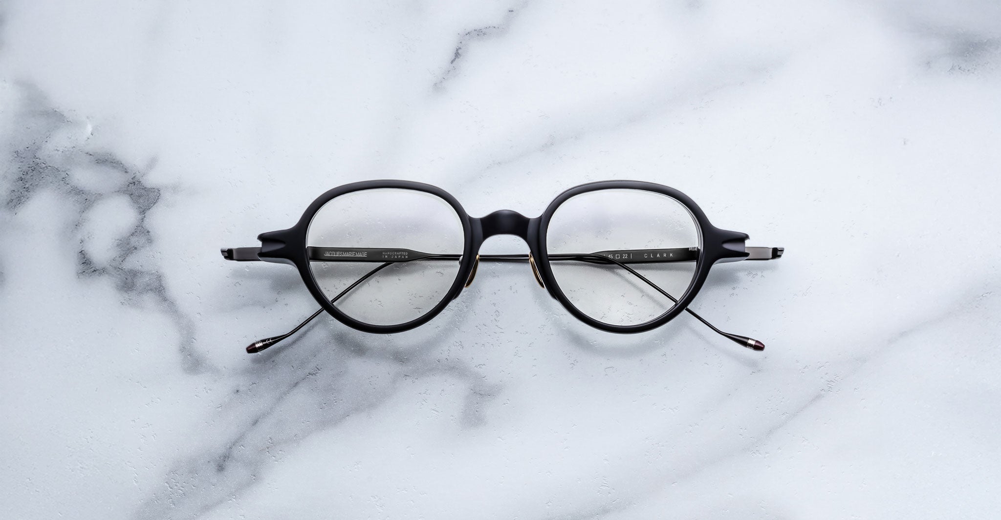 A pair of Jacques Marie Mage Clark round, black midcentury optical frames with thin temples is displayed on a white marble surface.