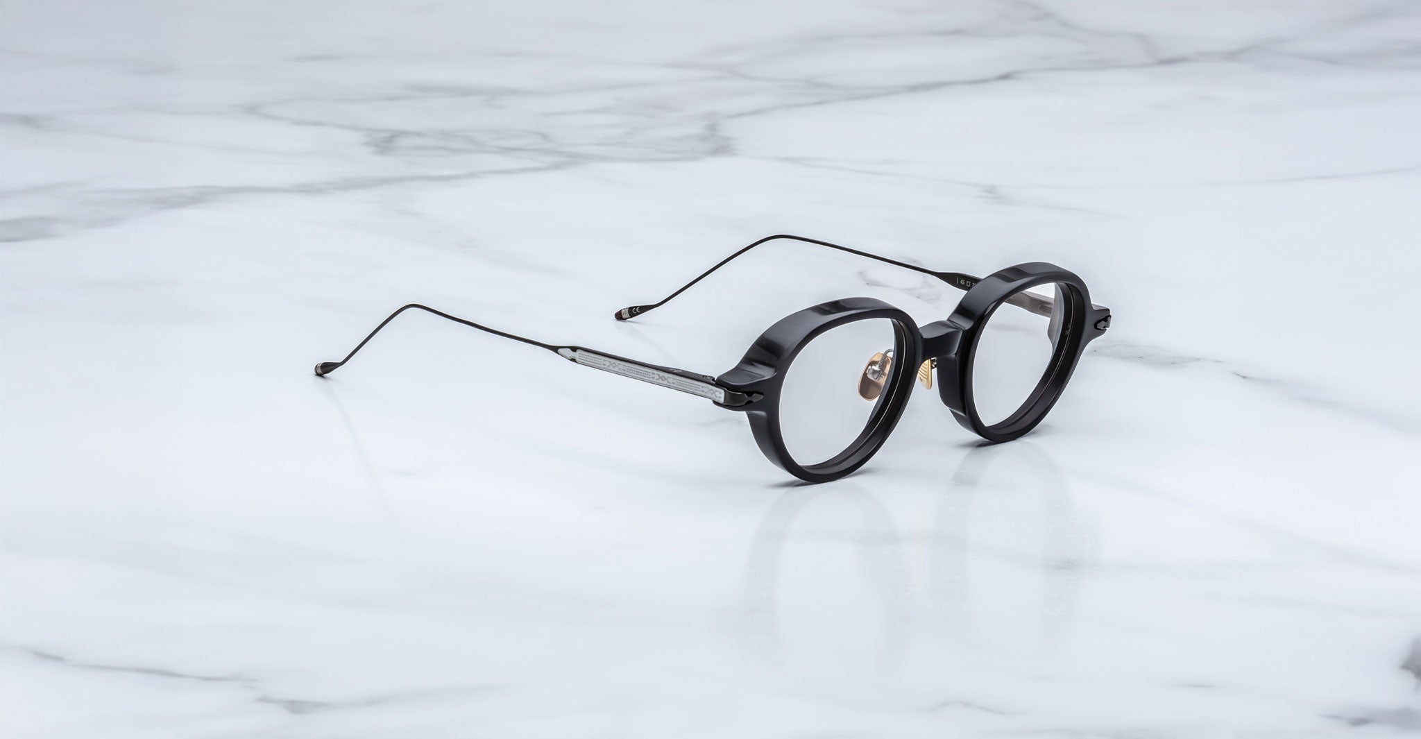 The Jacques Marie Mage Clark frames feature round black rims and thin metal arms, embodying midcentury-inspired design by Jacques Marie Mage. Shown elegantly on a white marble surface.