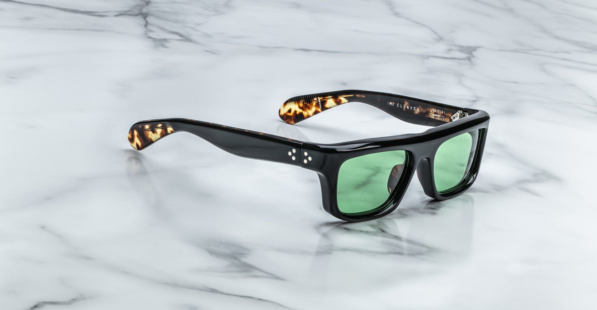 Jacques Marie Mage Cleavon sunglasses with black frames, green lenses, and tortoiseshell arms, shown from a top-down view.