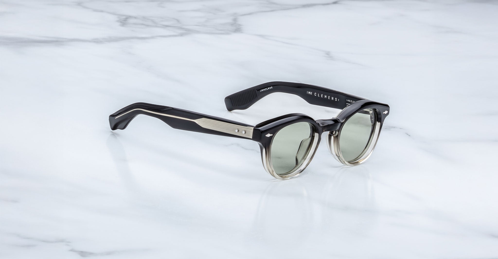 Jacques Marie Mage Clemens round sunglasses with green-tinted lenses and thick black-to-clear gradient frames rest on white marble. The black arms feature metallic accents and visible interior branding.