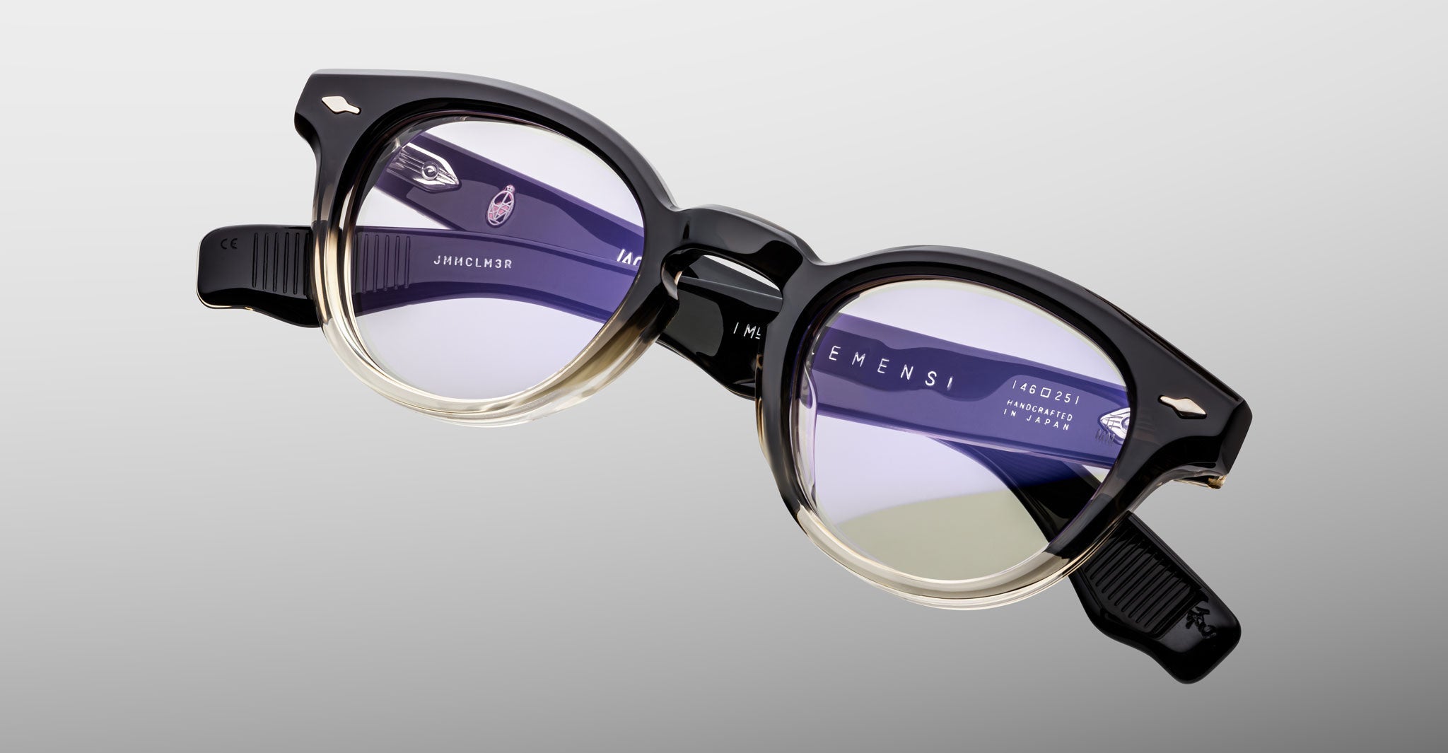 The Jacques Marie Mage Clemens eyeglasses by Jacques Marie Mage feature round black and clear frames with translucent lenses, shown on a plain gray background, embodying the brand’s signature refined craftsmanship.