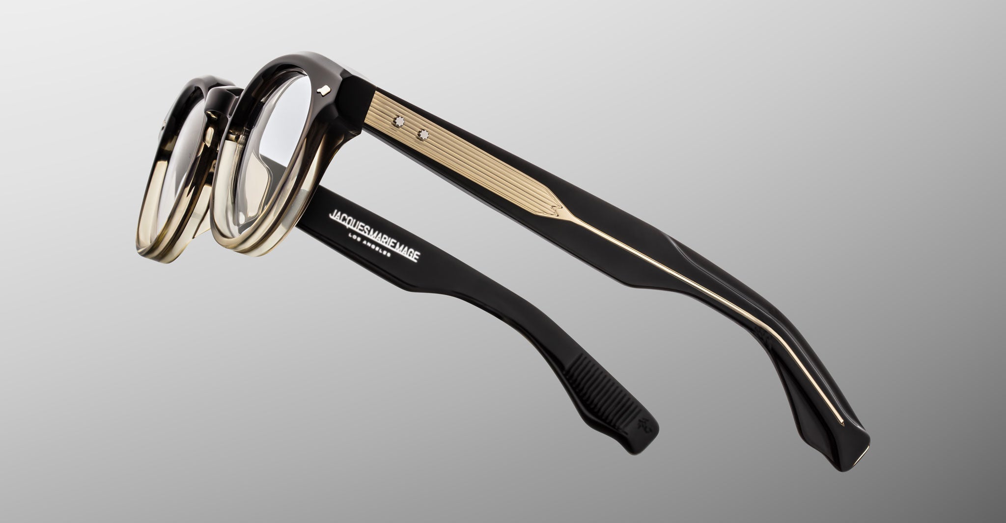 The Jacques Marie Mage Clemens by Jacques Marie Mage features black and gold thick frames with textured metal side accents, inspired by classic Wellington style, shown against a plain gray background.