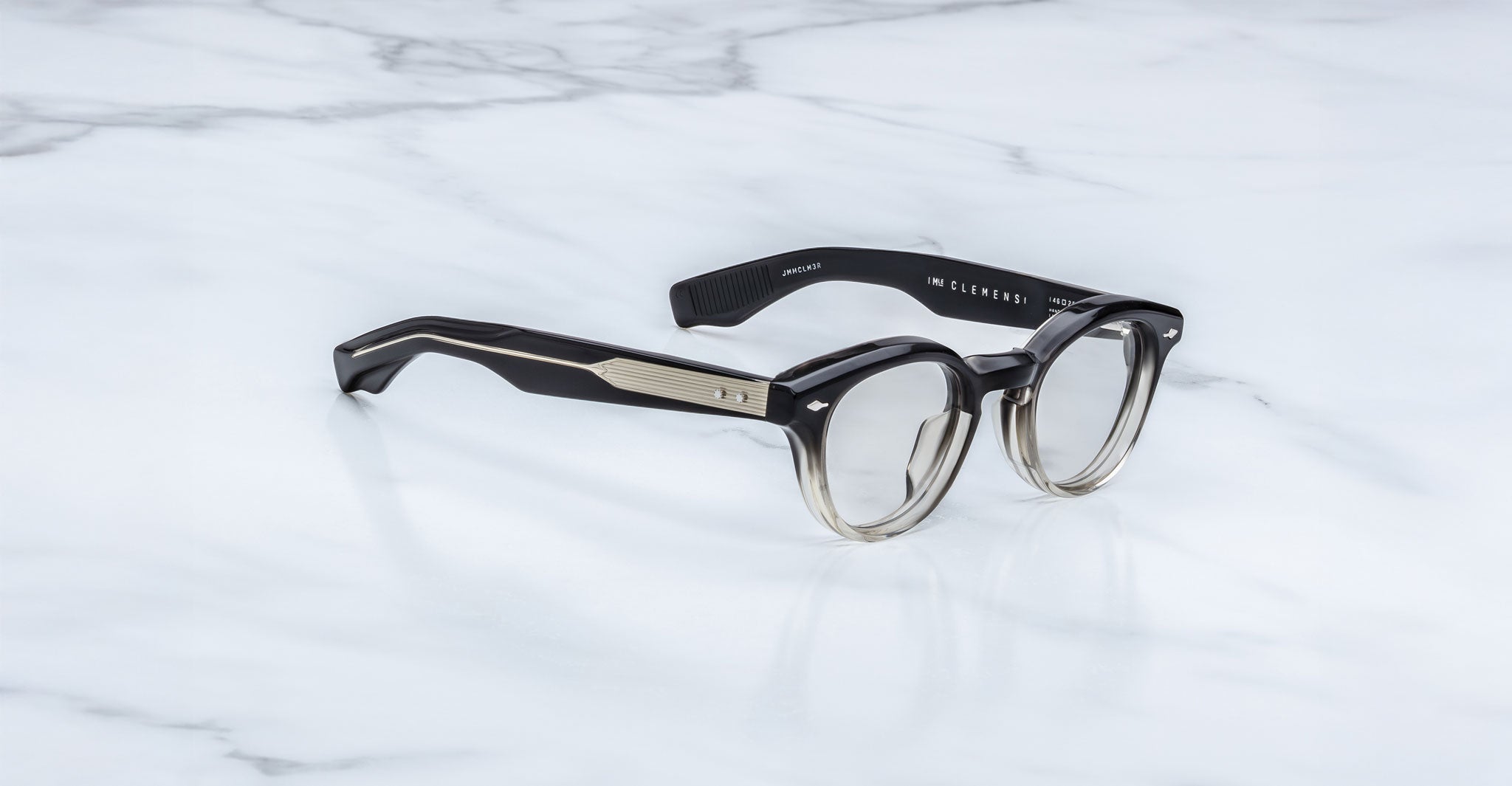 A pair of stylish, round Jacques Marie Mage Clemens eyeglasses with thick, black and translucent frames rest on white marble. The temples have metallic accents and the inside arm is labeled IN-ELEMENTS.
