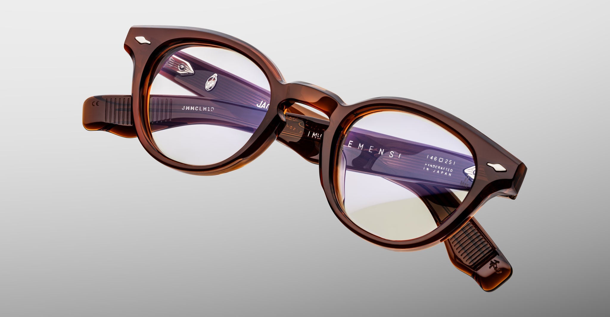 The Jacques Marie Mage Clemens eyeglasses feature a brown, round acetate frame inspired by Wellington design, with clear lenses, silver accents, and branded inner arms, all beautifully displayed against a neutral background.
