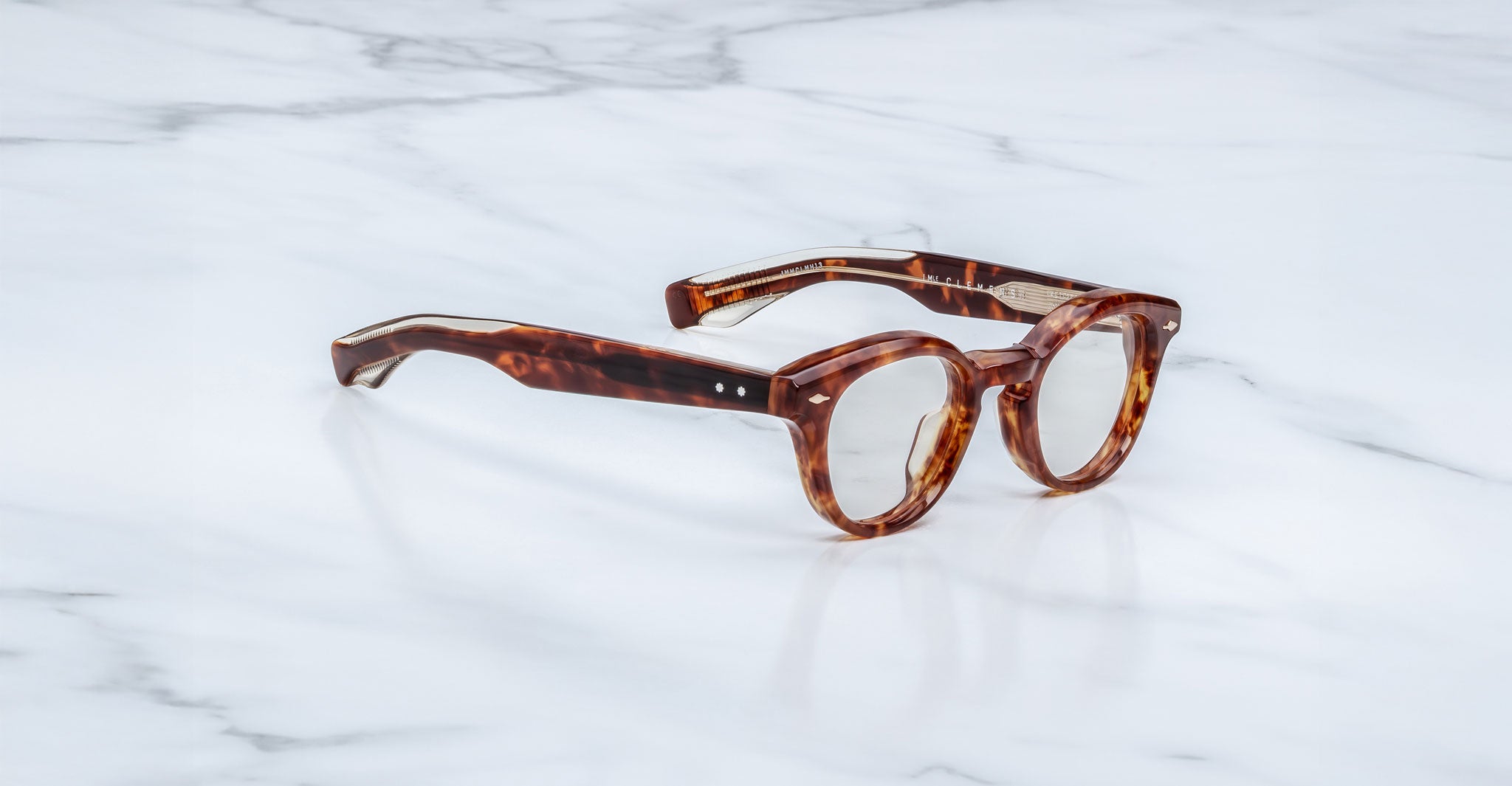 Jacques Marie Mage Clemens eyewear, featuring round lenses and thick brown tortoiseshell frames, rests on a white marble surface.