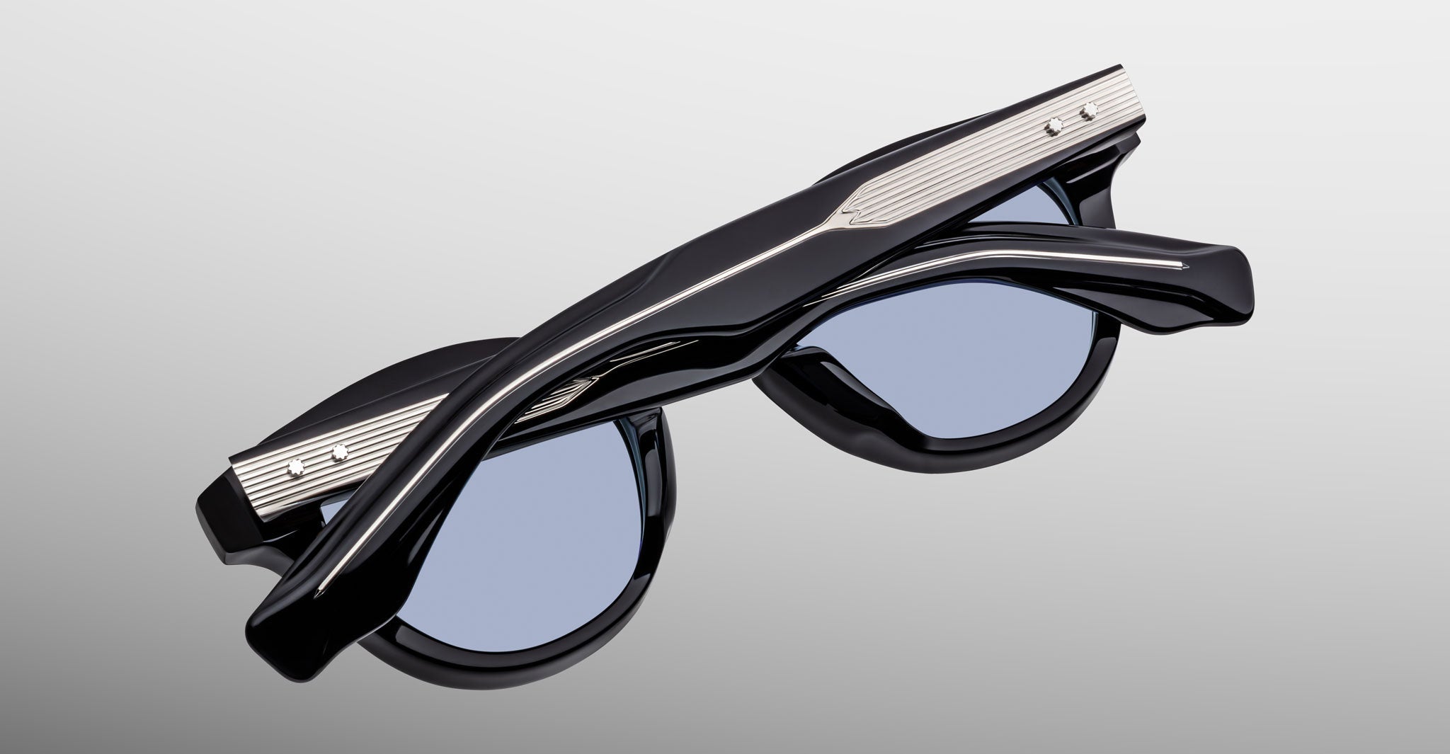 The Jacques Marie Mage Clemens sunglasses feature a folded black frame with blue-tinted lenses and metallic temple accents, presented on a light gradient background—a refined update to classic Wellington style.