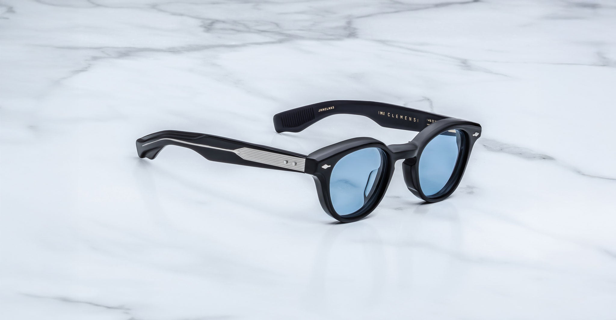 A pair of Jacques Marie Mage Clemens limited-edition sunglasses with black frames and blue-tinted lenses rests on white marble. The arms have metallic accents and gold writing inside.