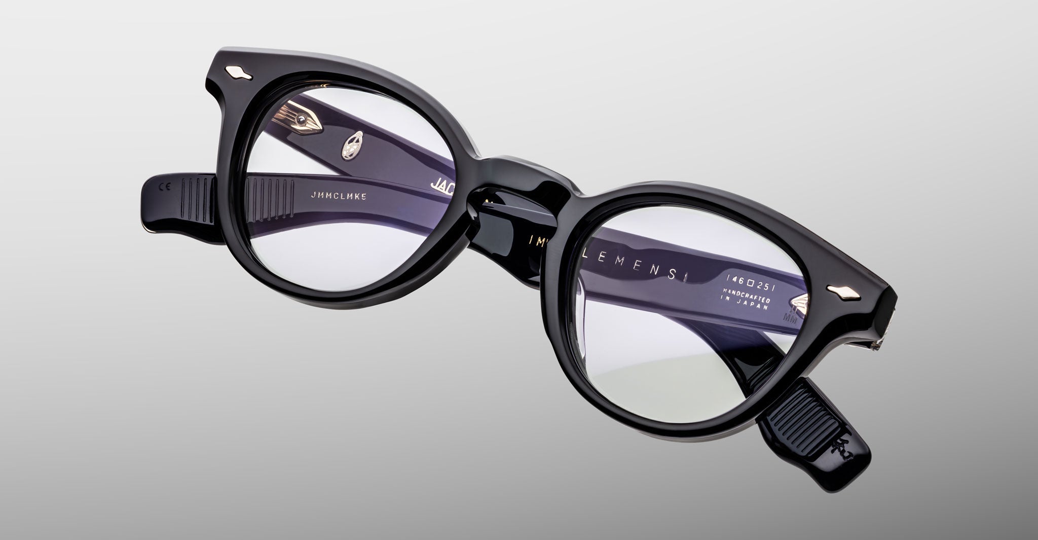 The Jacques Marie Mage Clemens eyeglasses by Jacques Marie Mage, featuring black round thick frames inspired by classic Wellington style, are displayed against a plain light gray background.