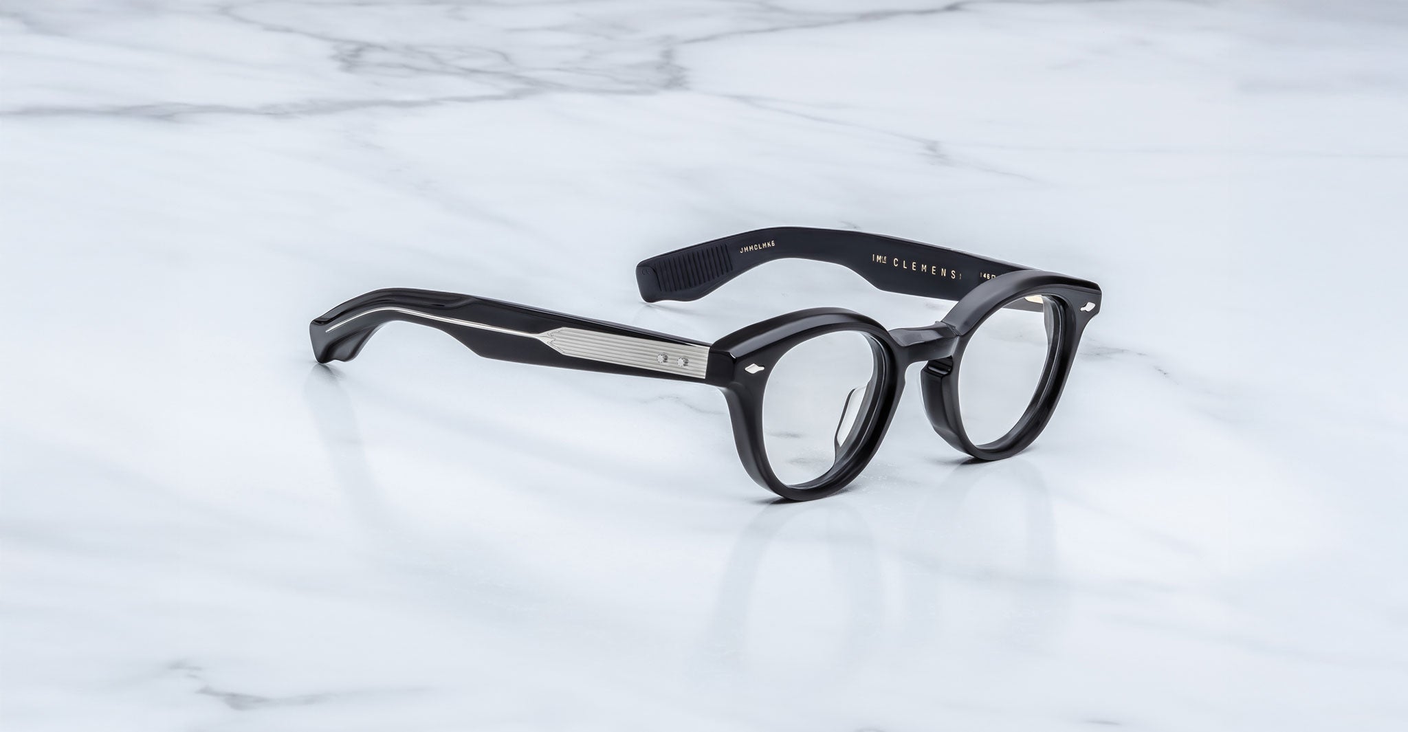 The Jacques Marie Mage Clemens features black round eyeglasses with thick frames and metal temple detailing, inspired by Wellington eyewear, displayed stylishly on a white marble surface.
